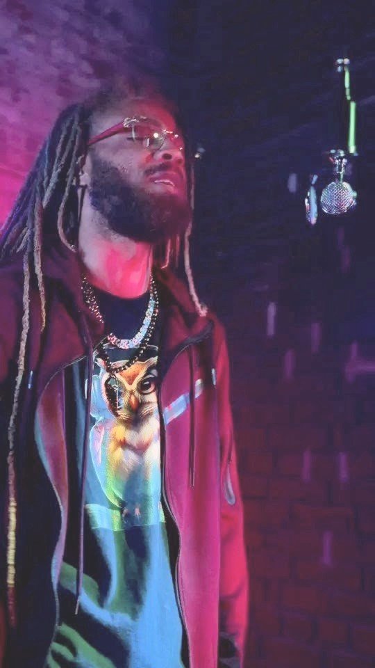 Person with long dreadlocks and glasses wearing a tiger graphic shirt and a jacket, standing near a microphone.