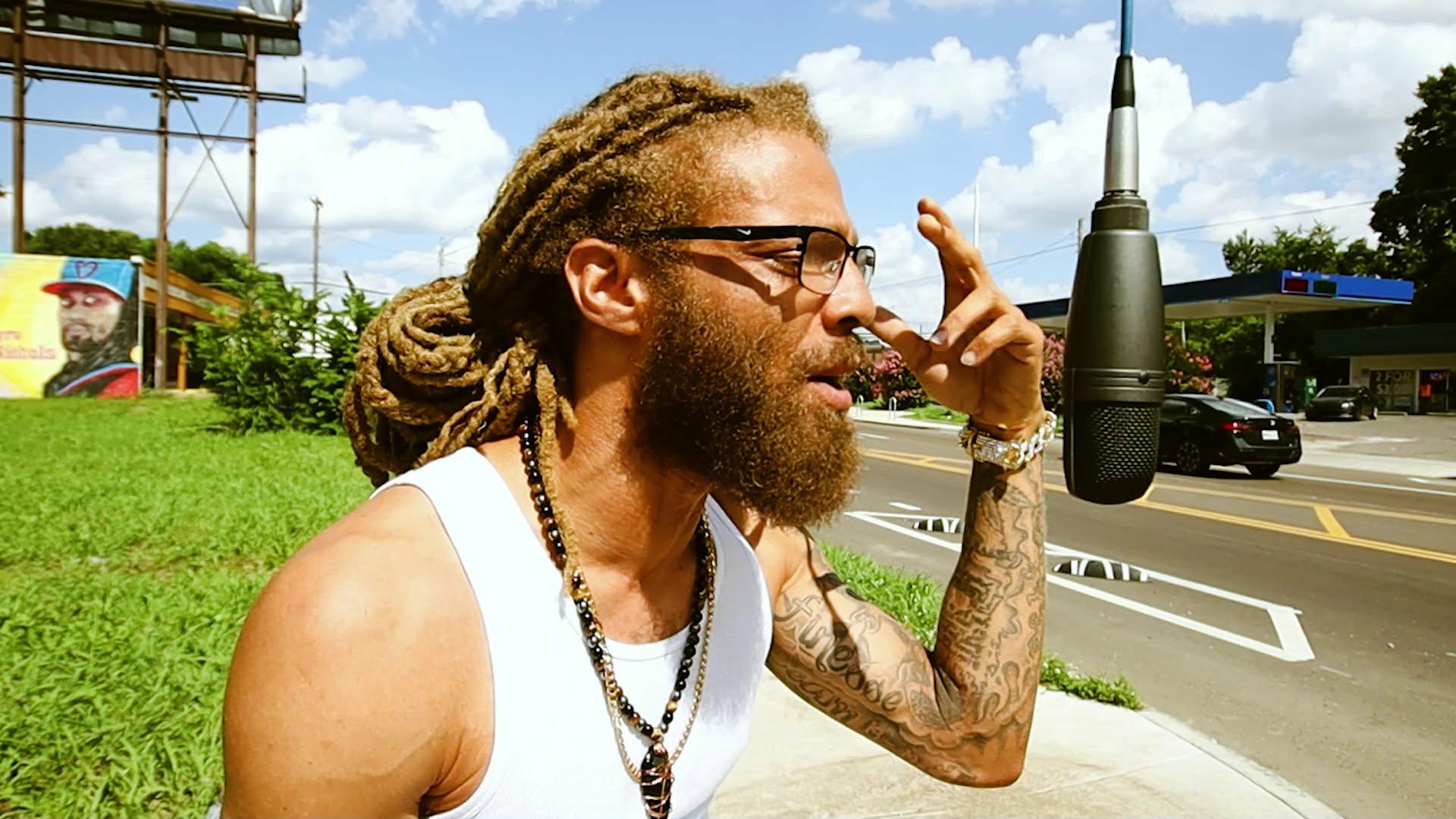 Man with dreadlocks and glasses singing into a microphone outdoors on a sunny day.