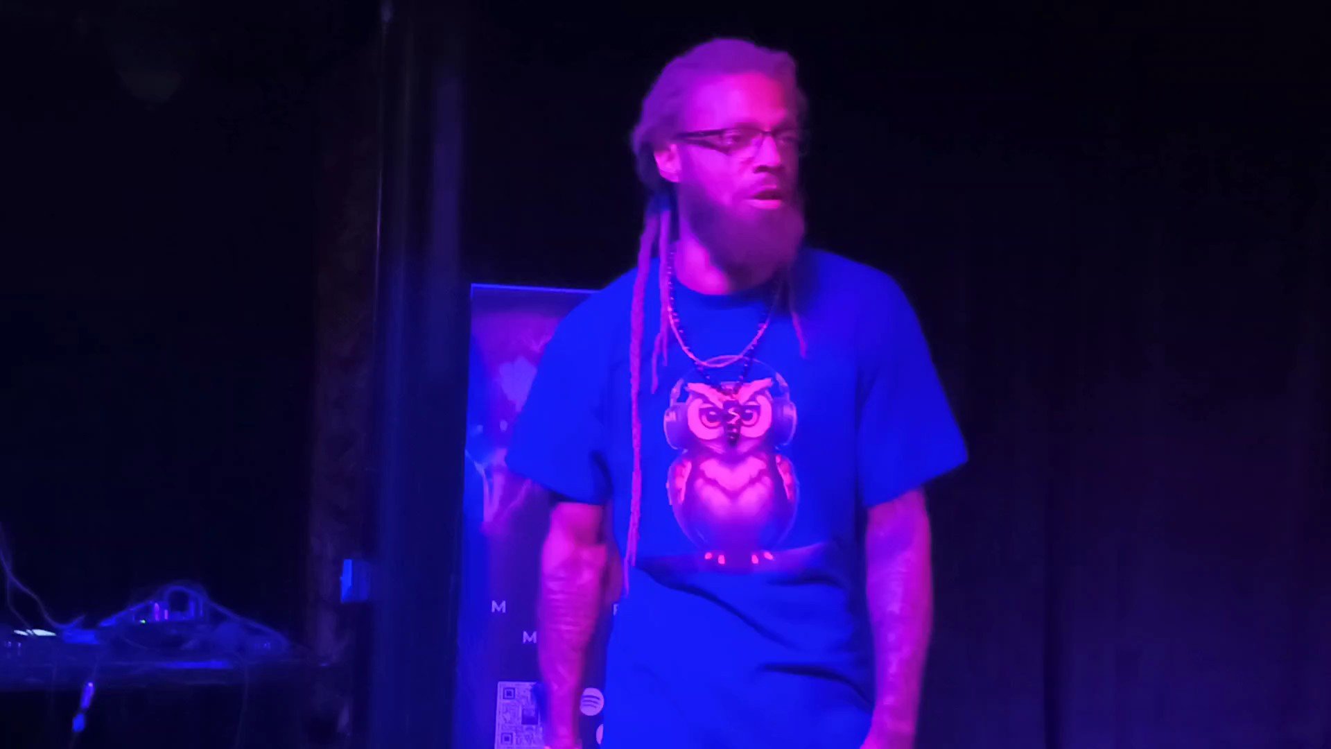 Person with long hair and glasses standing on stage in front of a dark background, wearing a T-shirt with a graphic design.