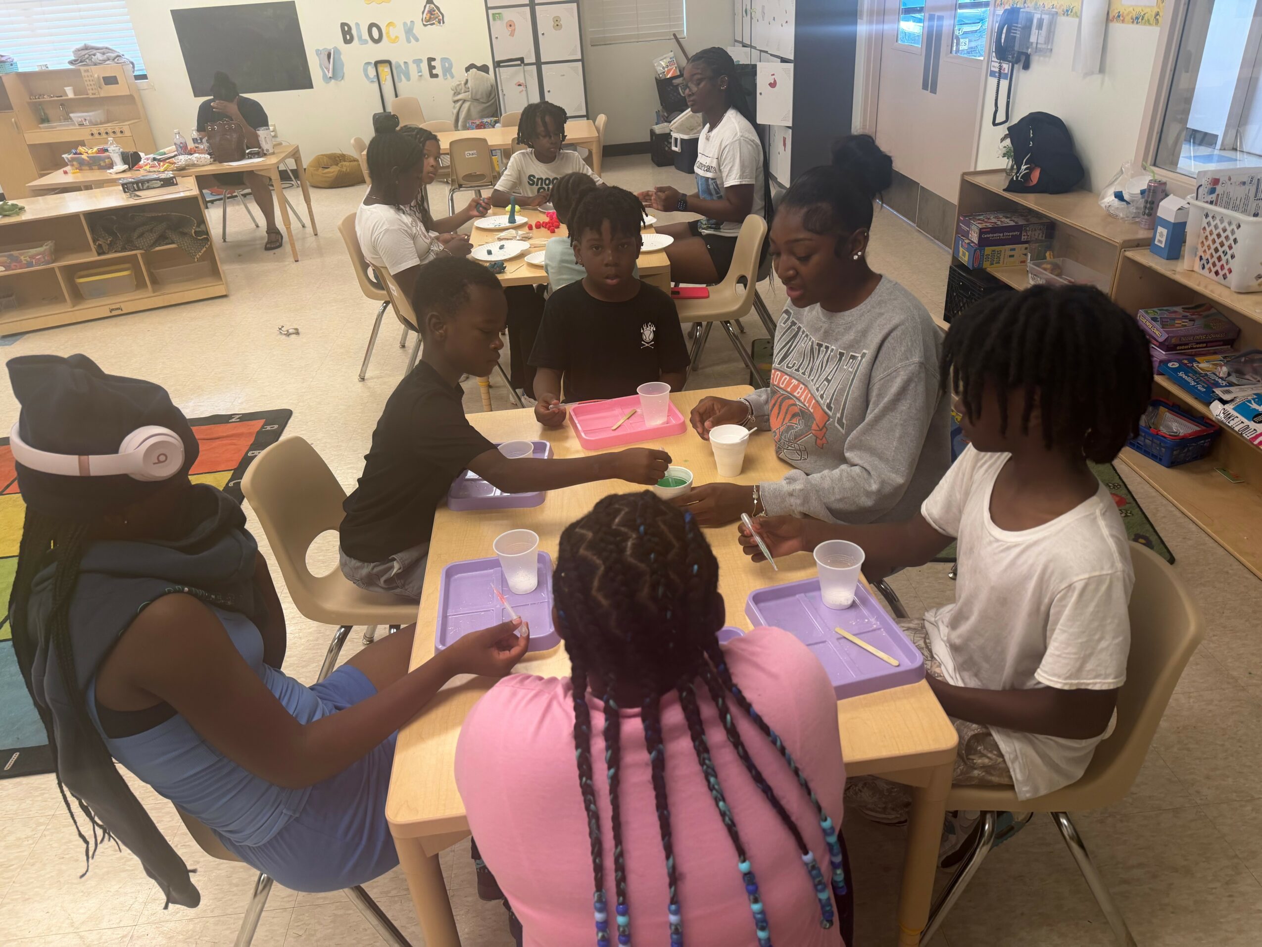 Children and adults sit around tables in a classroom, engaging in an activity with trays and cups, with bookshelves and windows in background.