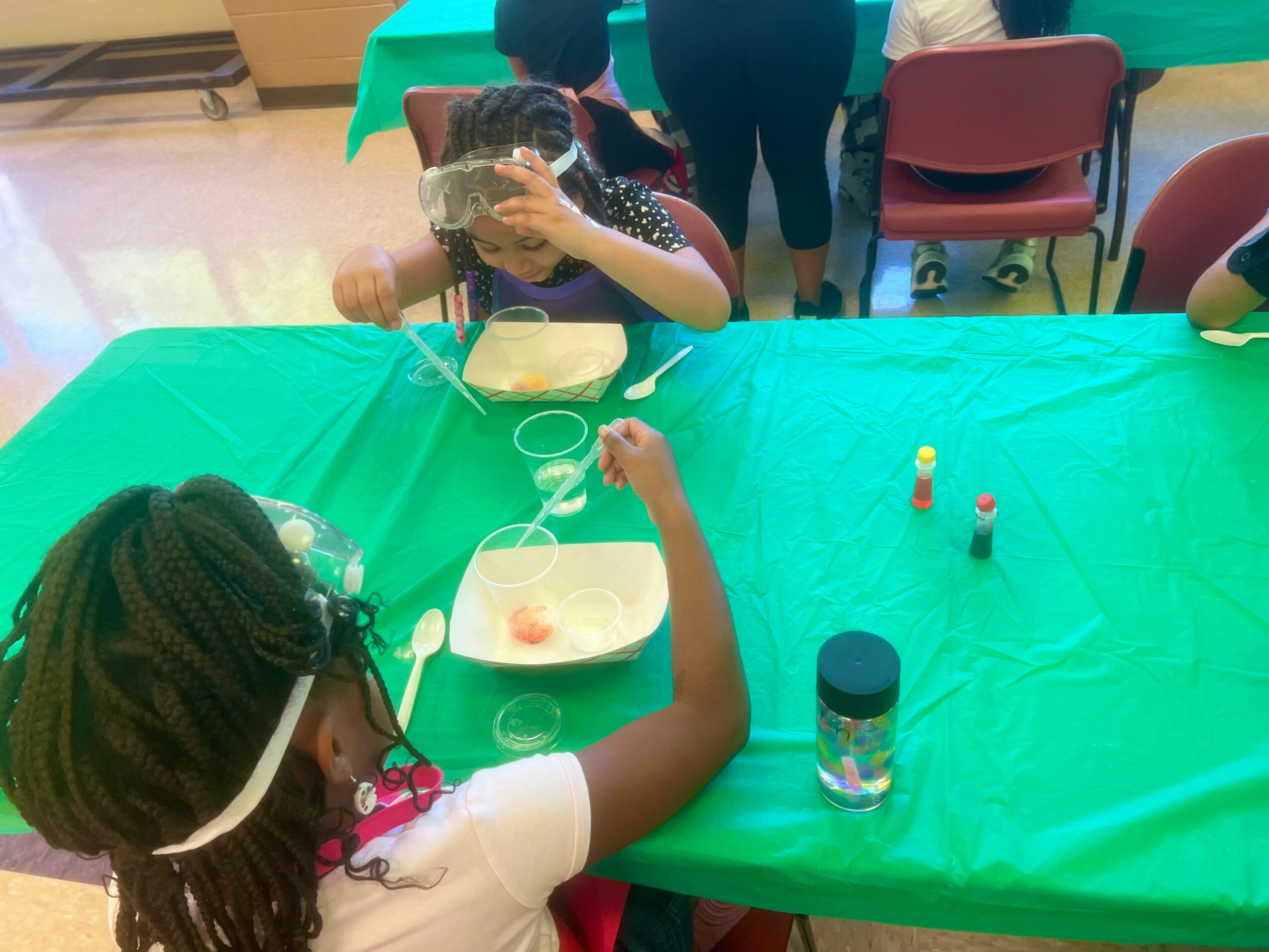Child with braided hair and glasses at a green table, holding a spoon, with a water bottle and small objects nearby.
