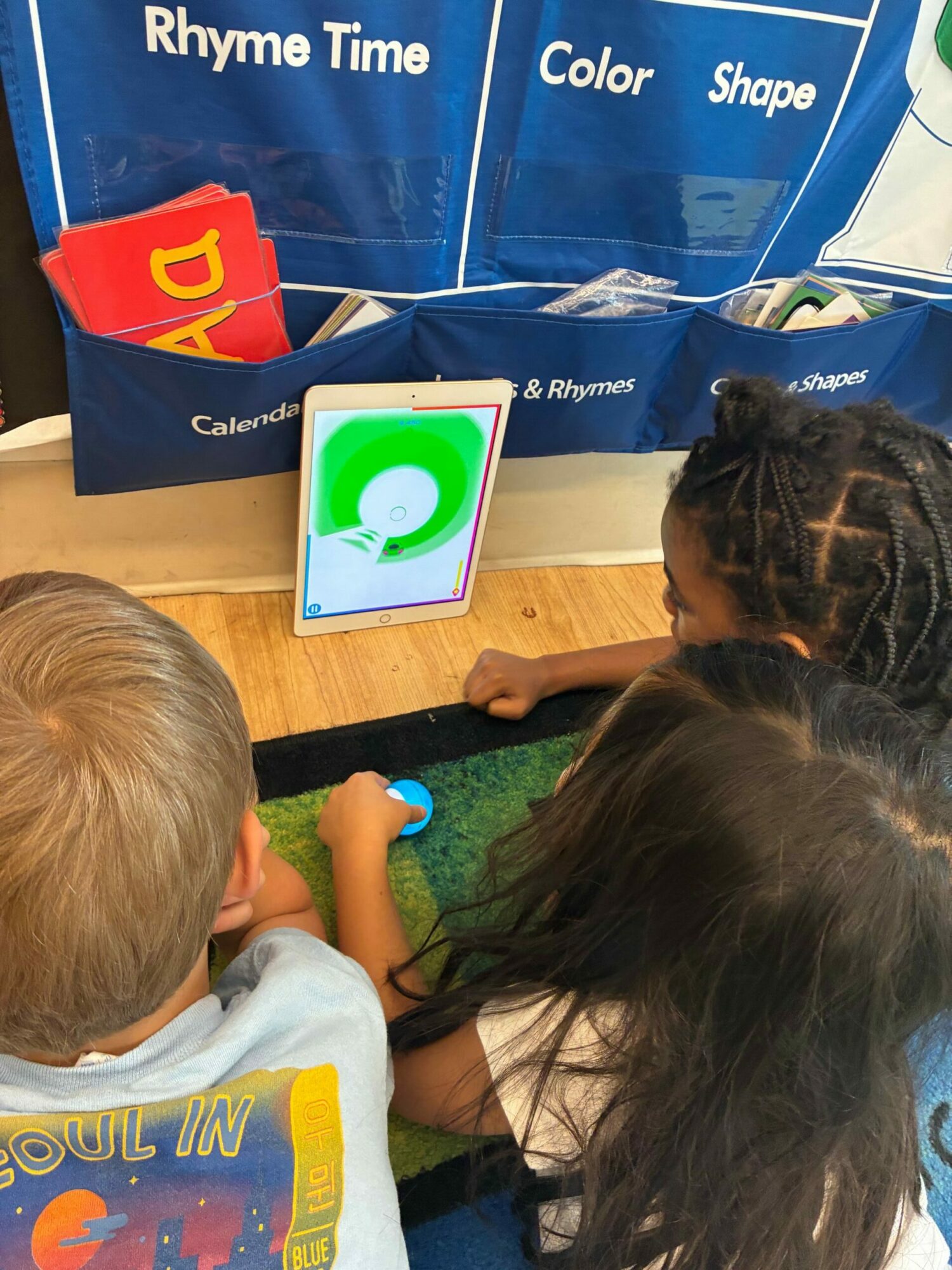 Children gather around a tablet displaying a colorful circular chart, with educational materials nearby.