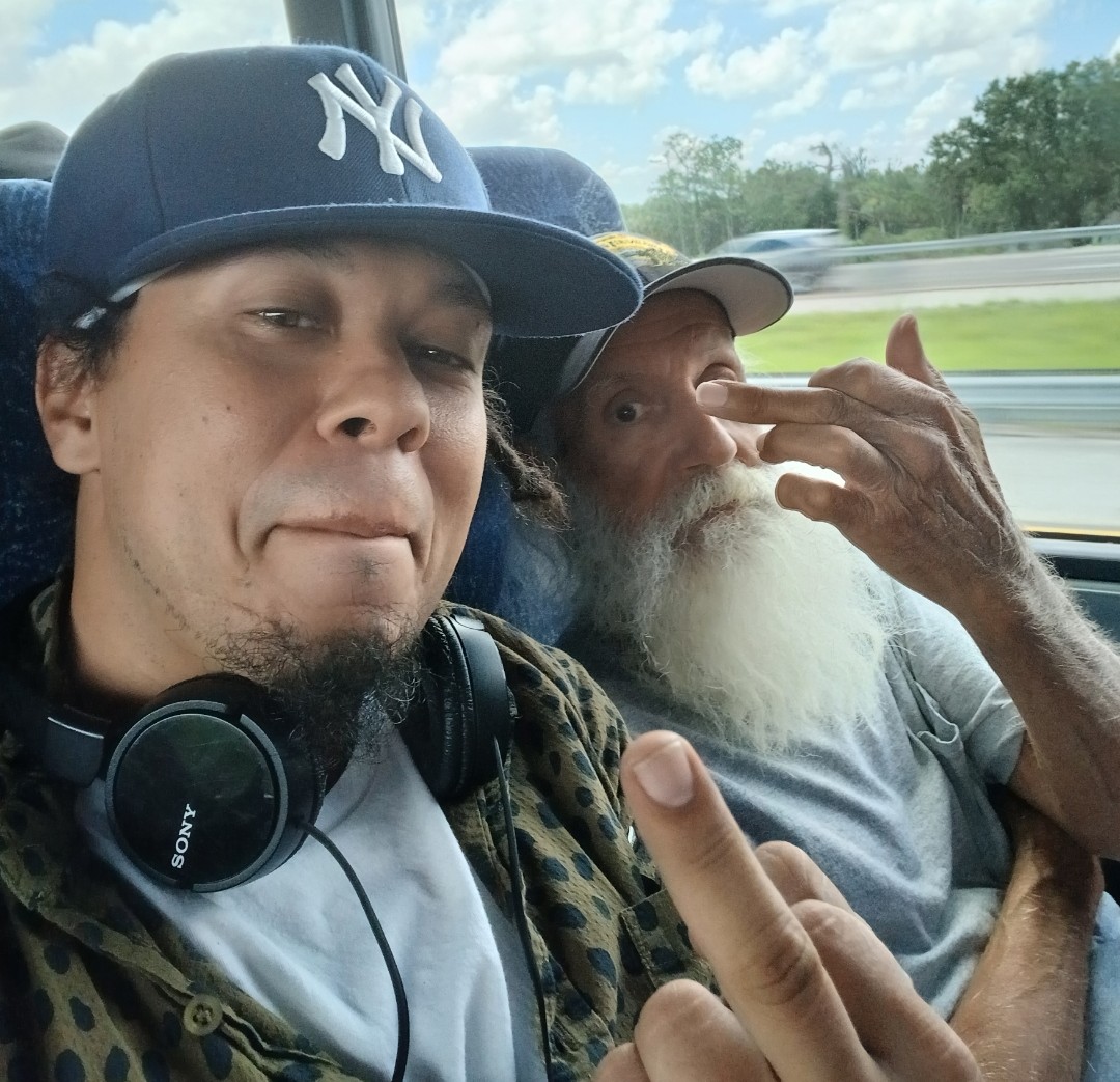 Two men sitting on a bus, one wearing a baseball cap and headphones, the other with a beard, making gestures.