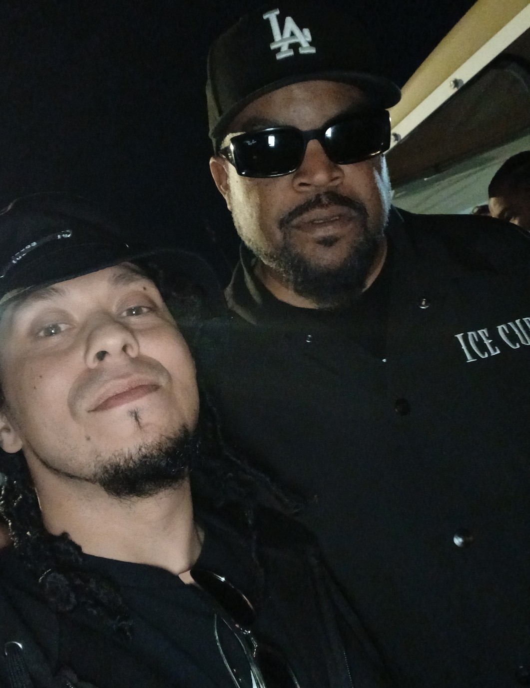 Two men pose for a photo, one wearing sunglasses and a cap, the other with a goatee and a cap, at night.