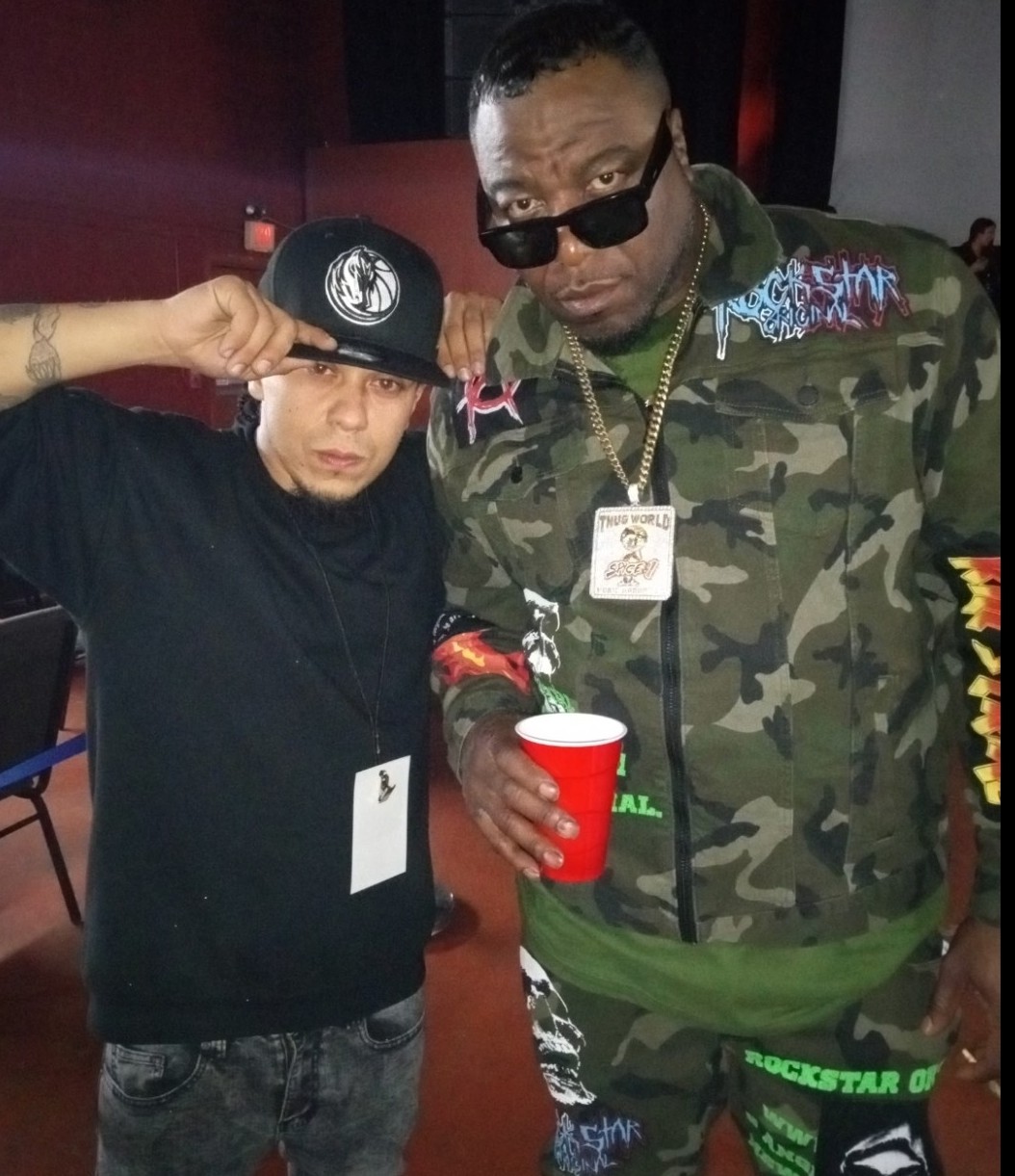 Two men pose indoors, one wearing sunglasses and a camouflage jacket, holding a red cup; the other adjusts a cap.
