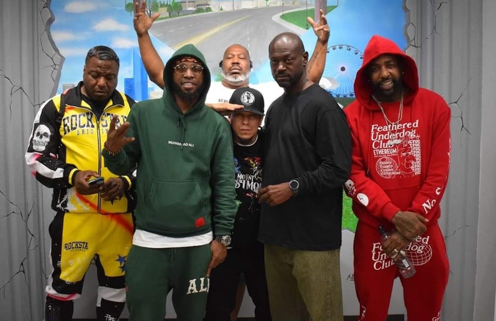 Group of six people standing indoors, some with raised hands, colorful background, casual clothing, and hoodies.