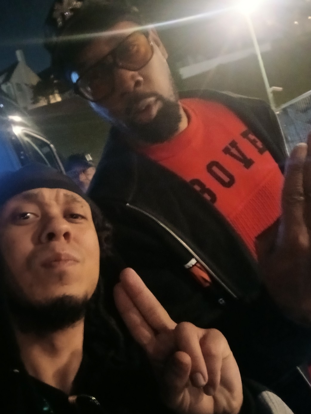 Two men posing outdoors at night, one making a peace sign, illuminated by streetlights.
