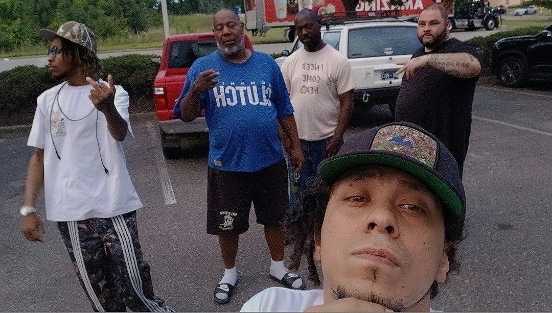 Group of five men standing outdoors in a parking lot, one taking a selfie, some with tattoos and casual clothing.