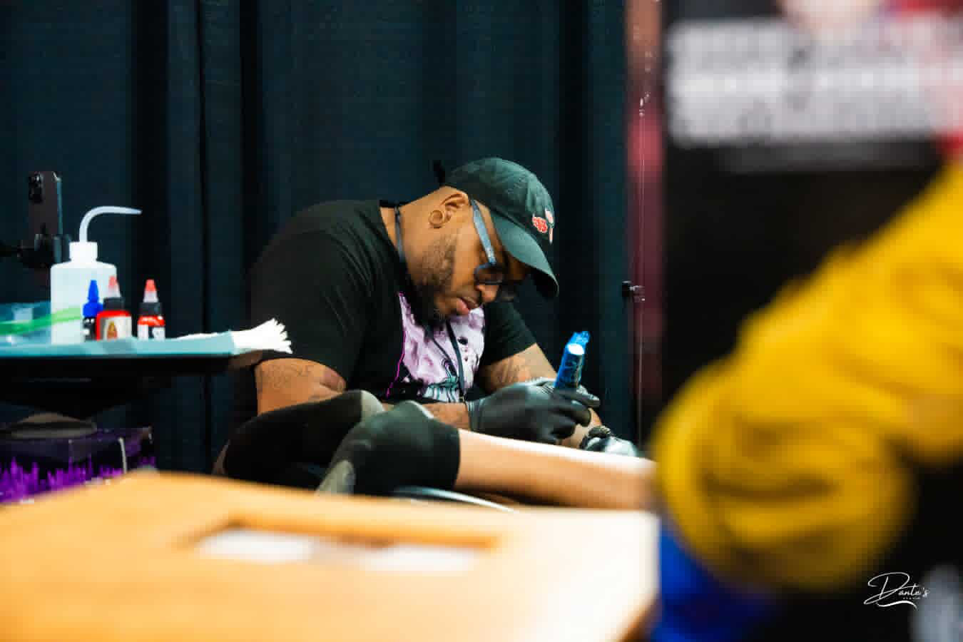 Tattoo artist working on a client's arm in a tattoo studio, with tattoo supplies nearby, and a person in yellow in the foreground.