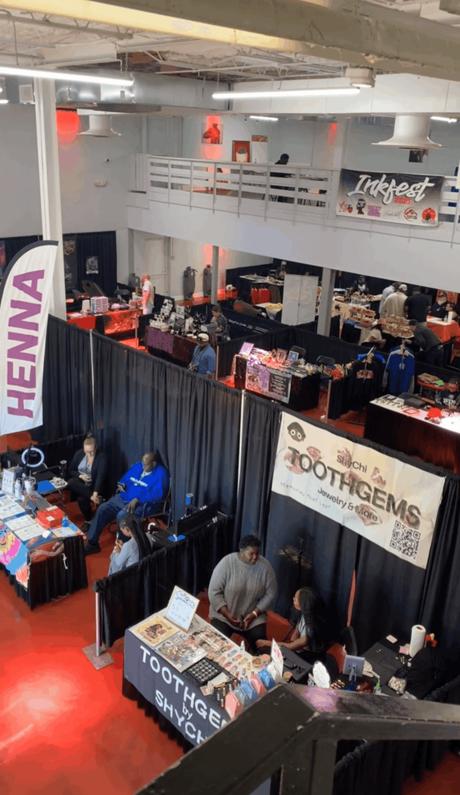 Indoor event with booths and people, banners display 'HENNA' and 'TOUR IMAGES', upper level with railing, ceiling lights.