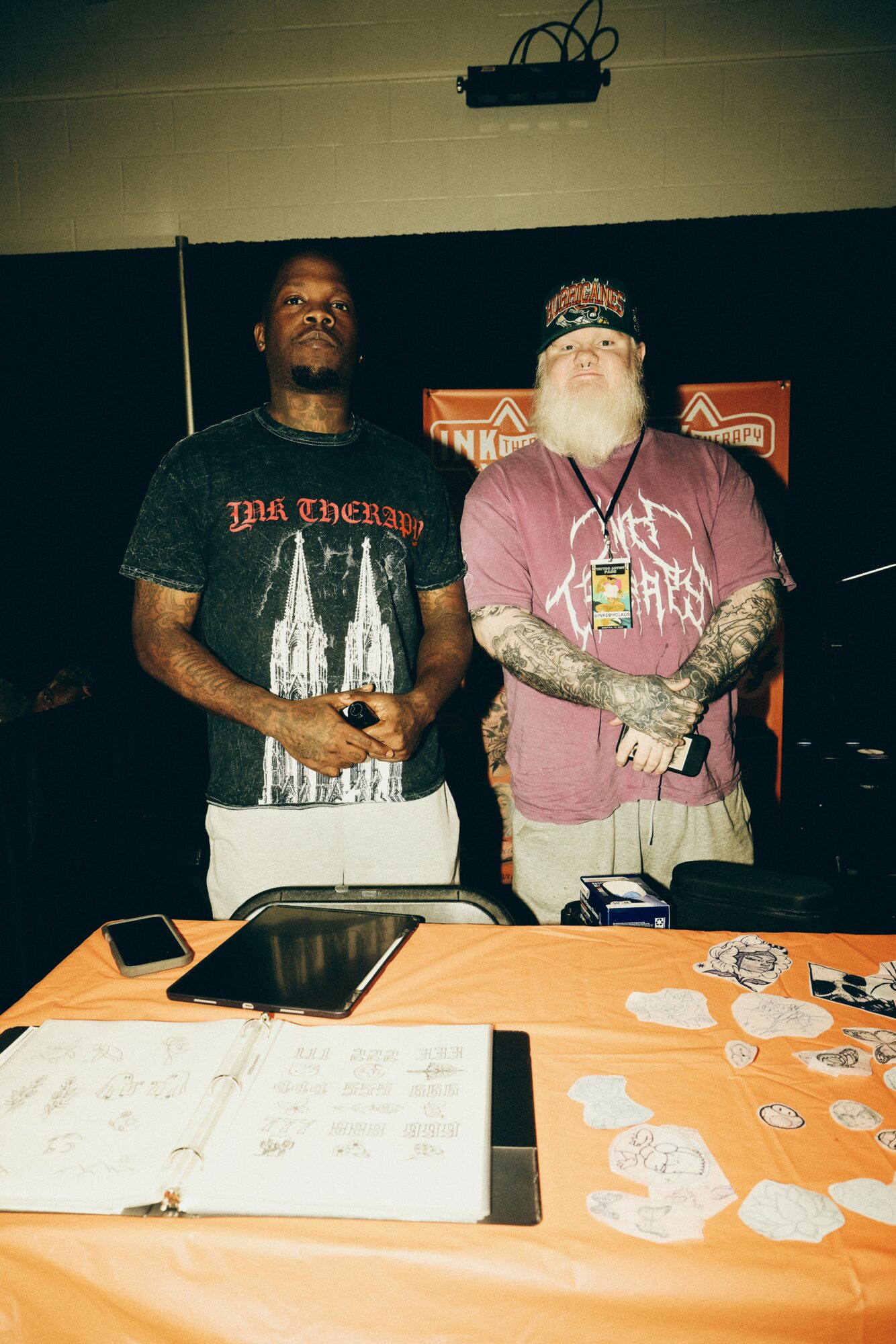 Two men standing behind a table with papers and items, one wearing a black T-shirt and the other a pink shirt with tattoos and a beard.