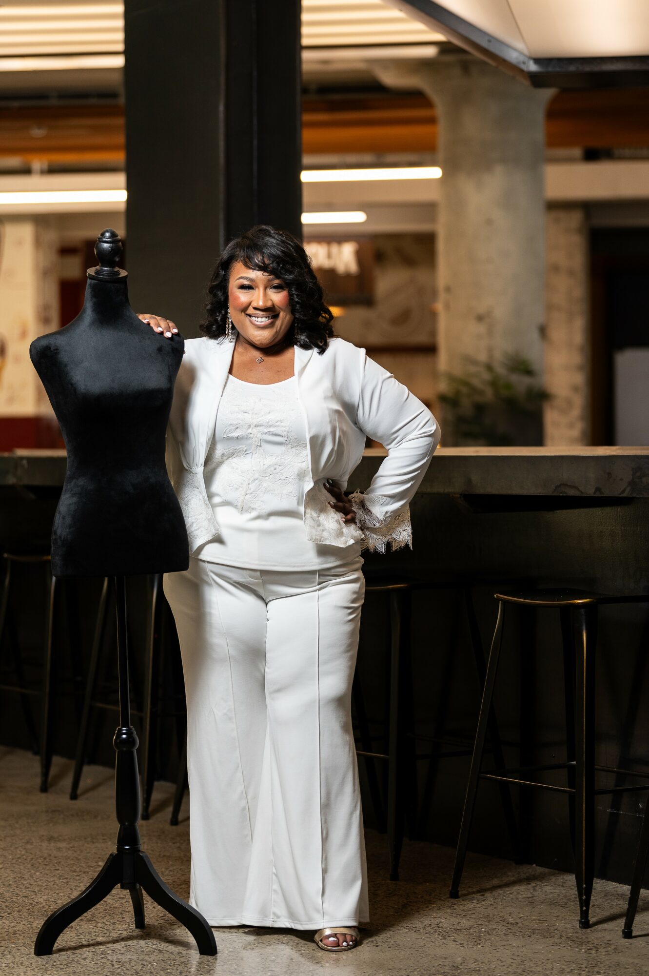 Woman in white outfit standing next to a dress form in an industrial-style space, smiling, with bar stools nearby.