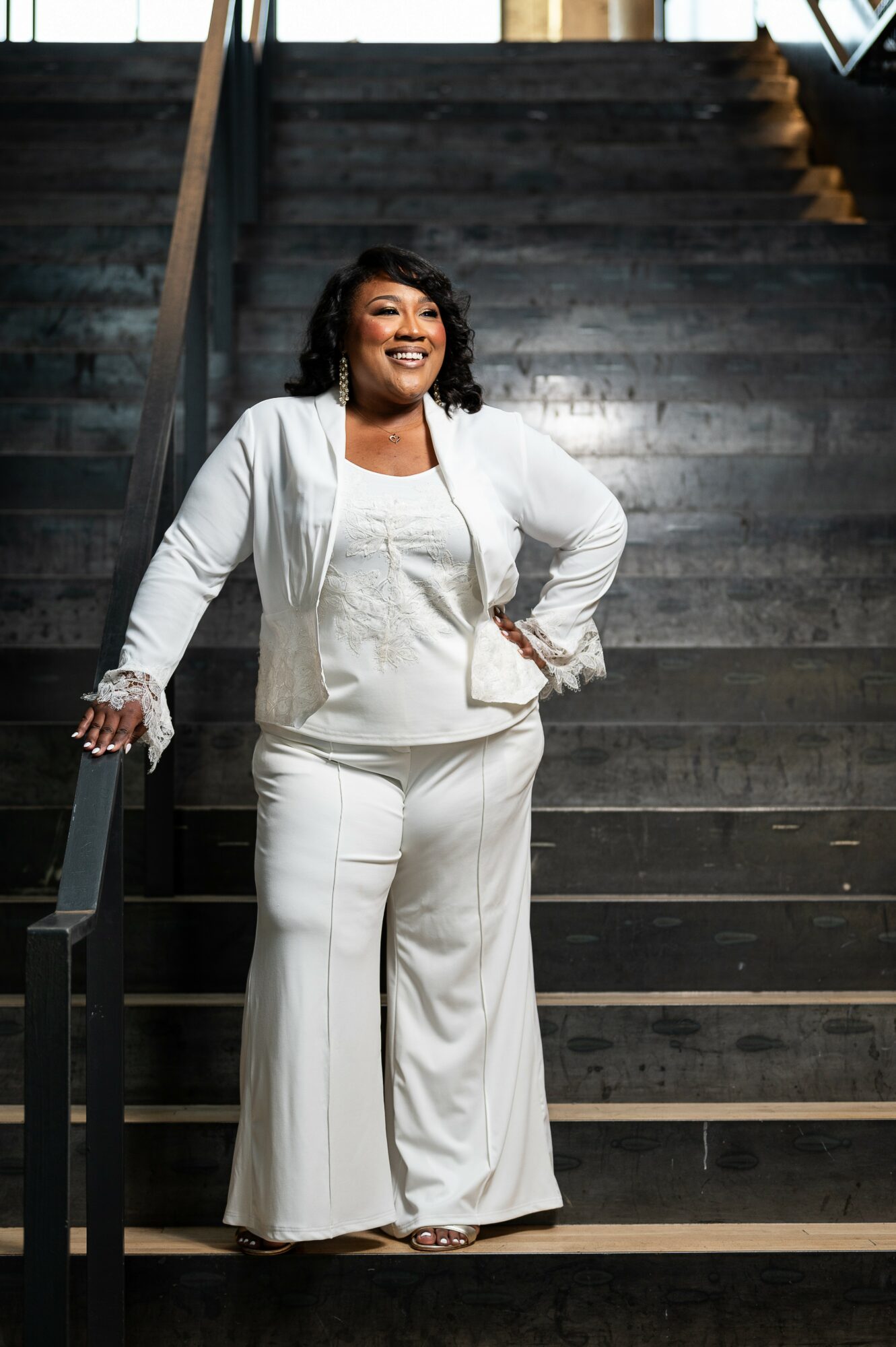 Woman in white outfit standing on stairs, smiling, with hand on hip, in an indoor setting.