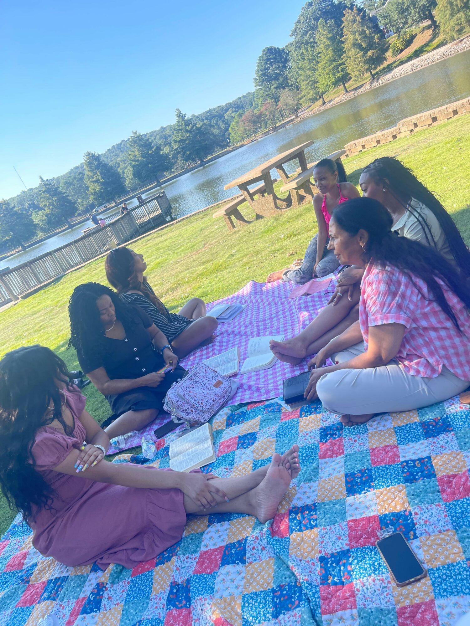 Two women sitting on a colorful quilt outdoors near a lake, with trees and a fence in the background.
