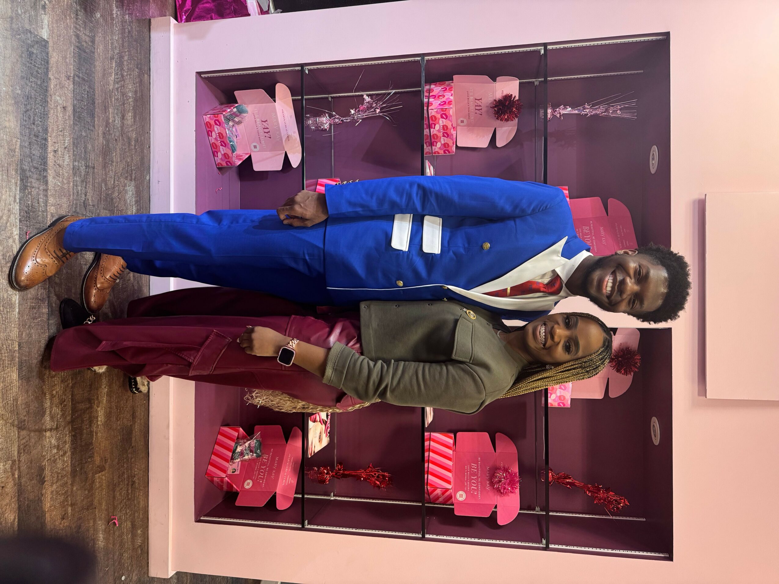 Two people standing in front of pink shelves with gift bags, one in a green blazer and the other in a blue suit.