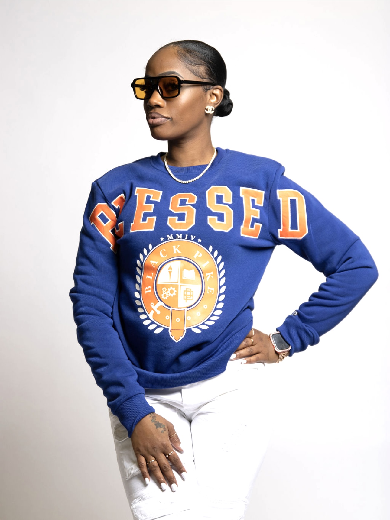Woman wearing sunglasses and a blue sweatshirt with the word 'BESSED' and a graphic, standing with hand on hip against a white background.