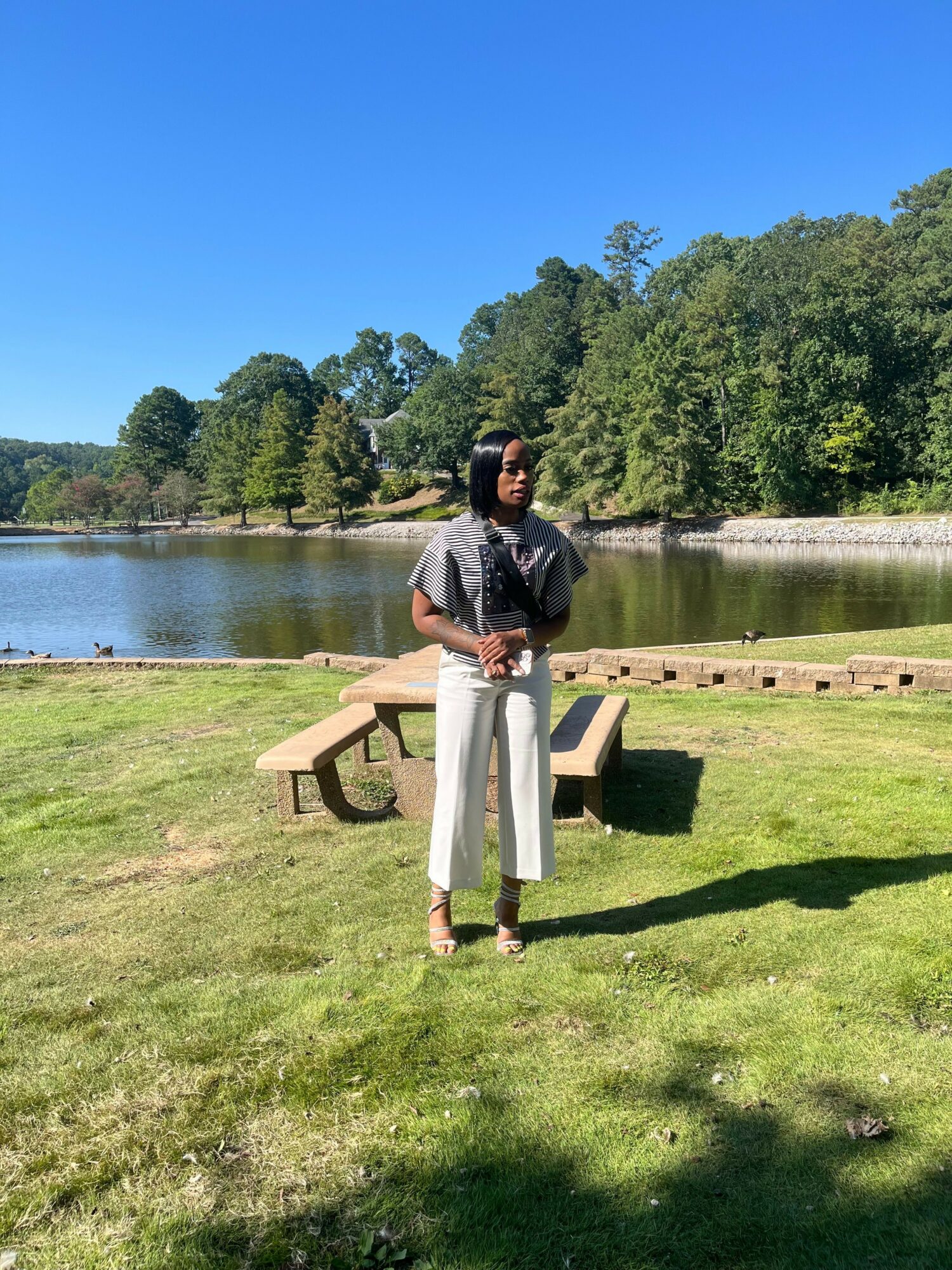 Person standing on grass near a pond with trees and a clear blue sky in the background.