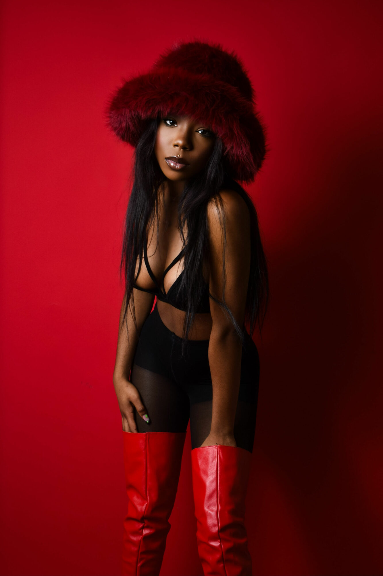 Woman with long dark hair wearing a red furry hat, black top, and red thigh-high boots against red background.