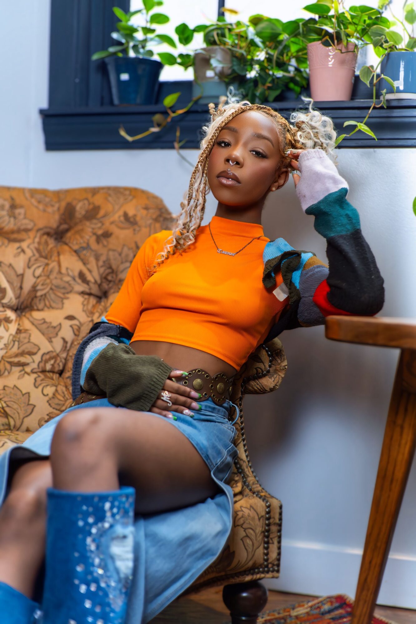 Young woman with blonde braids sitting on a patterned armchair, surrounded by potted plants, wearing an orange top and denim shorts.