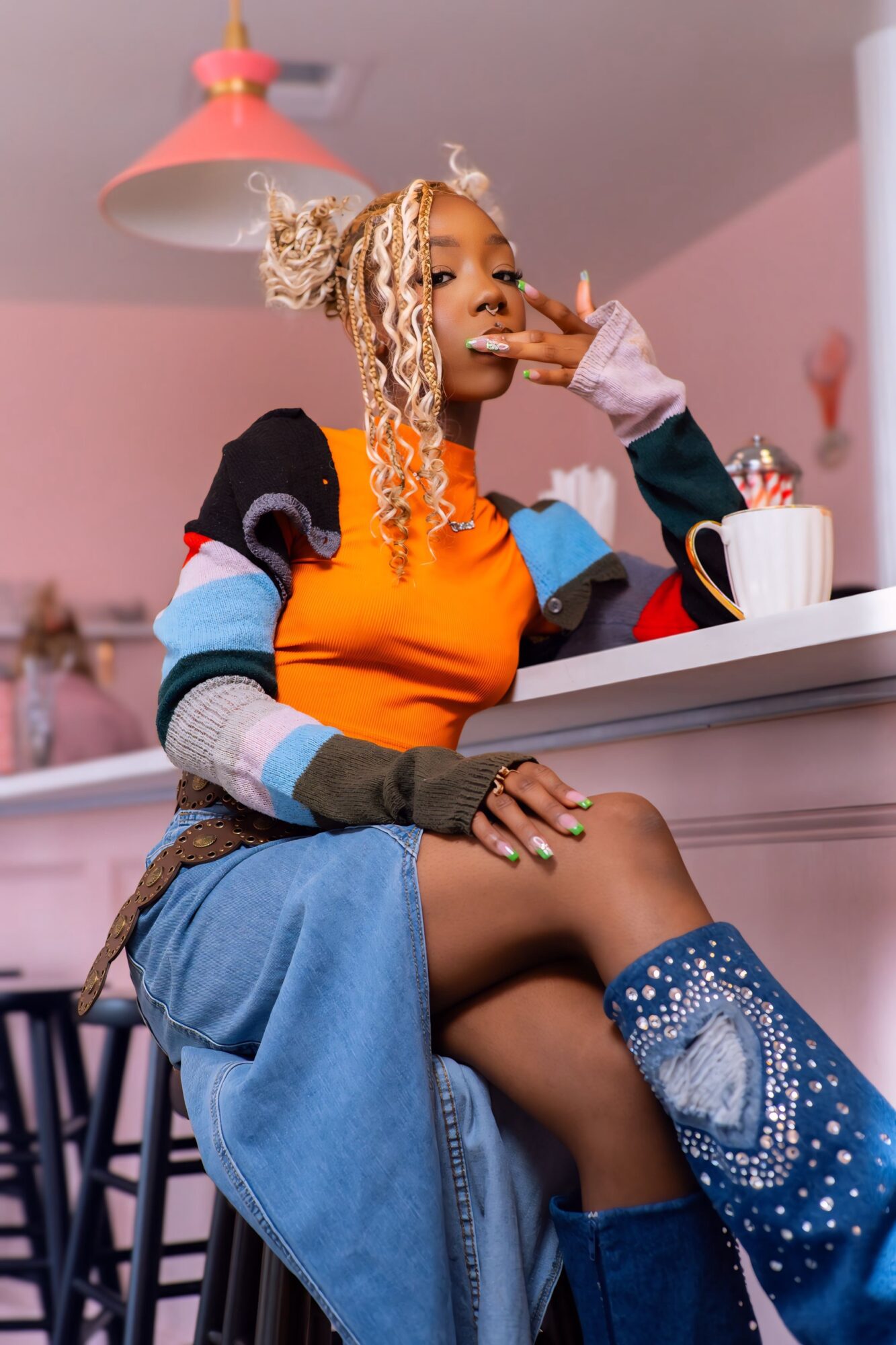 Young woman with blonde curly hair sitting at a table in a pink room, wearing colorful clothing and blue boots, with a mug nearby.