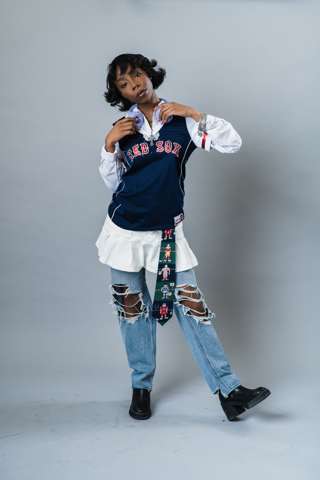 Young woman with curly hair wearing a layered outfit, standing against a plain background.