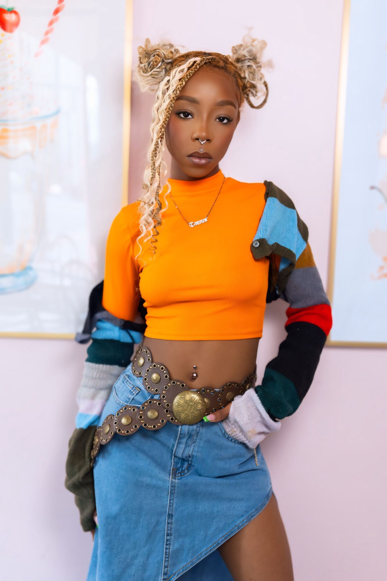 Young woman with blonde hair styled in two buns, wearing an orange crop top and a colorful patchwork jacket, standing indoors.