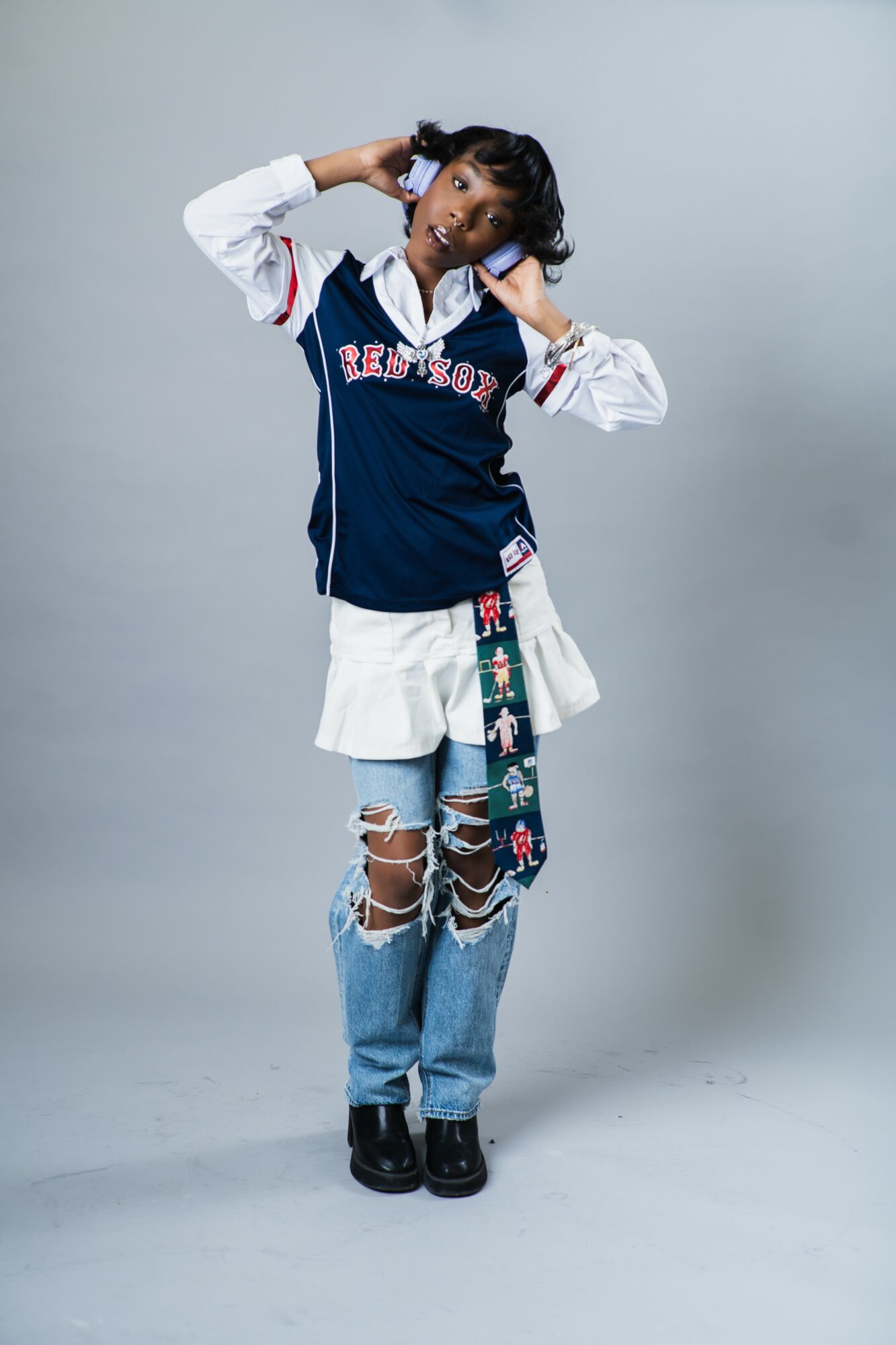 Young woman with curly hair wearing a baseball jersey, ripped jeans, and sneakers, standing against a plain background.