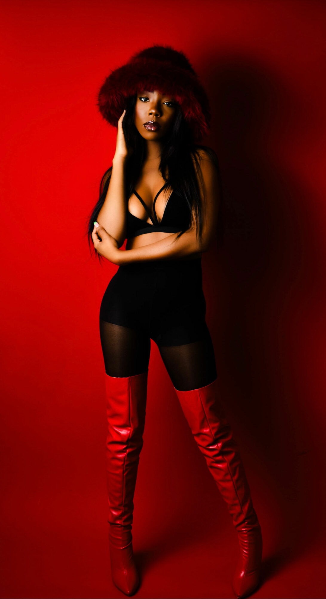 Woman with curly hair in black outfit and red thigh-high boots stands against red background.