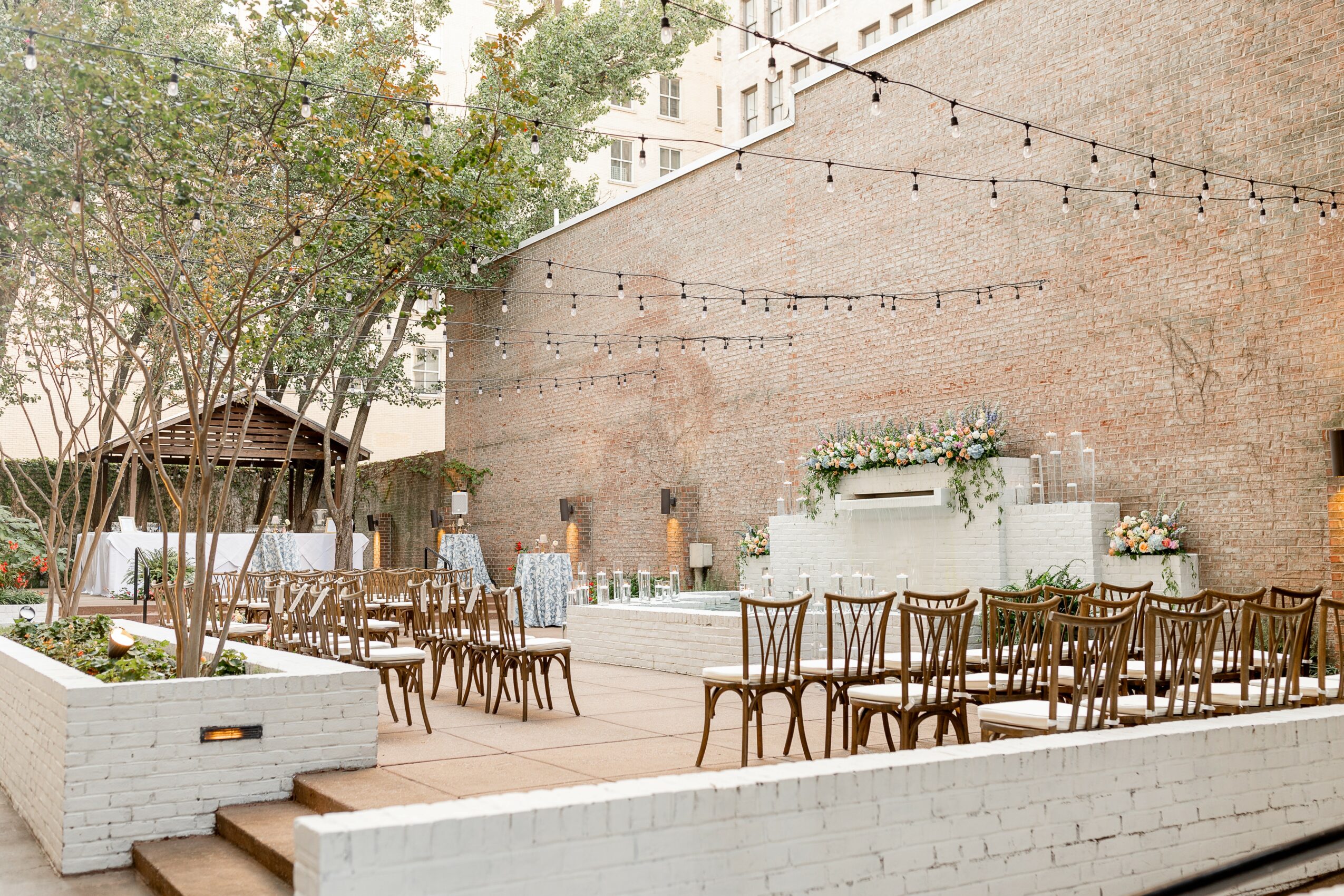 Outdoor patio with tables and chairs, string lights, trees, and a brick wall background.
