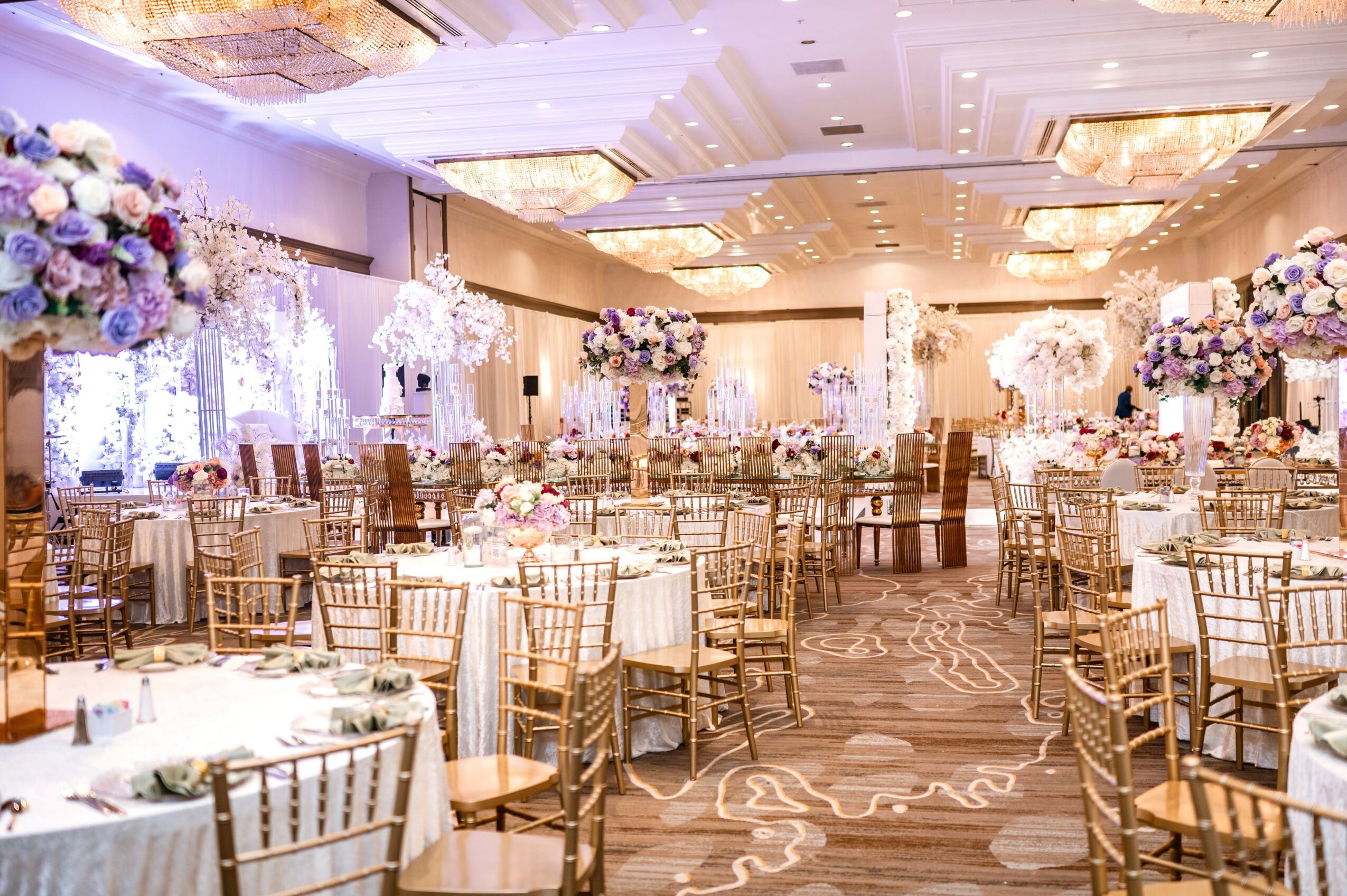 Elegant banquet hall decorated with floral centerpieces, round tables, and gold chairs, illuminated by chandeliers and soft lighting.