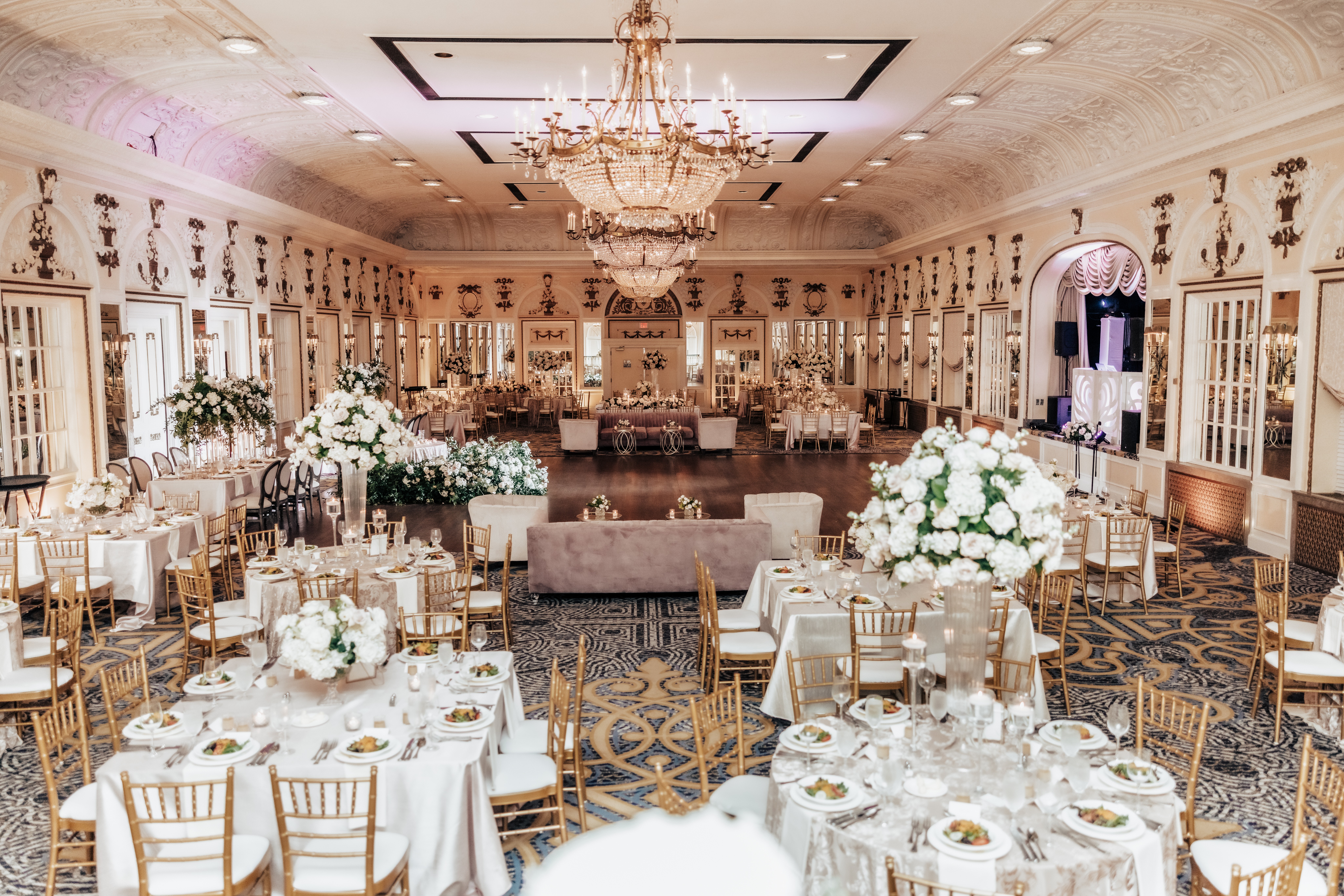 Elegant banquet hall with chandeliers, decorated tables with white flowers, and gold chairs, ready for a formal event.