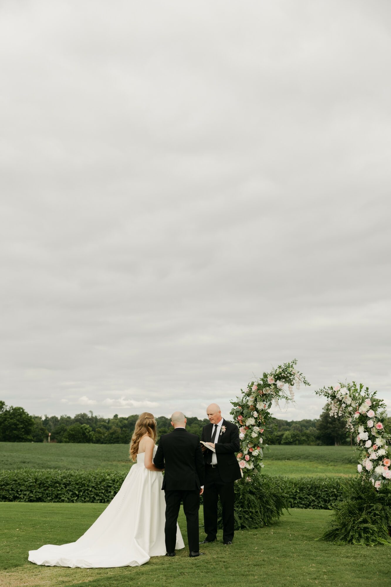 A wedding ceremony outdoors with a bride, groom, and officiant standing under an arch of flowers on a grassy field.
