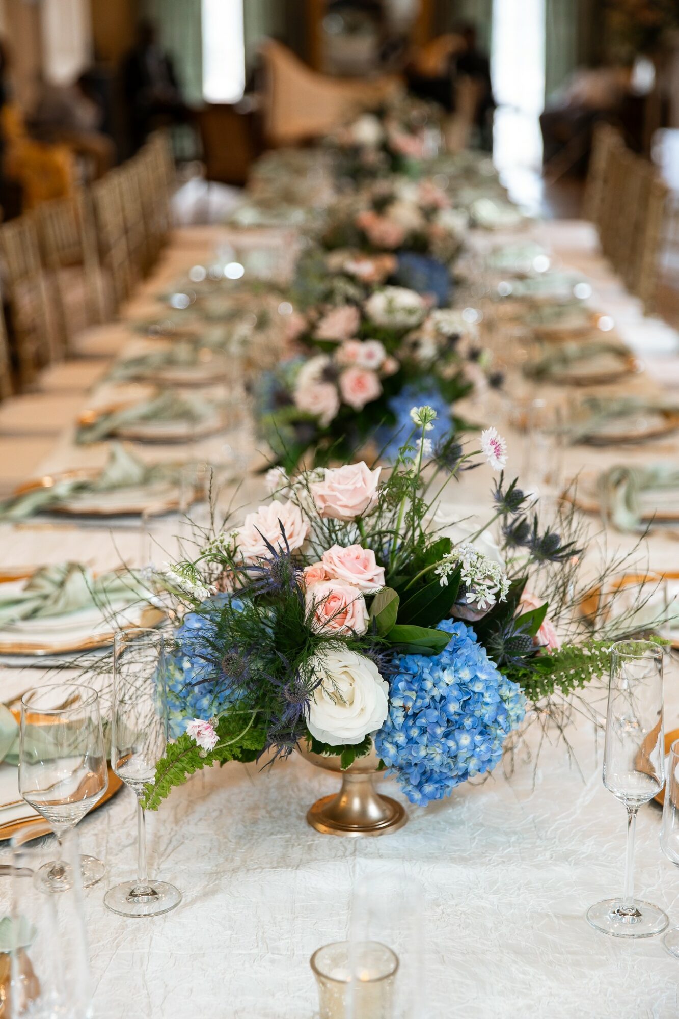 Long banquet table with floral centerpieces, set with plates, glasses, and cutlery, in a decorated indoor venue.