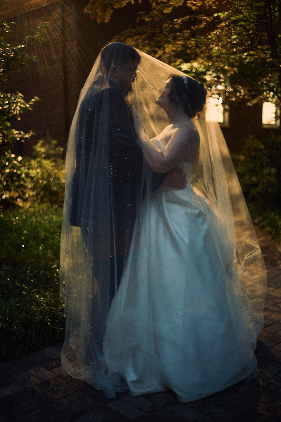 A bride and groom stand close together outdoors, with sunlight filtering through trees, sharing a moment under a veil.