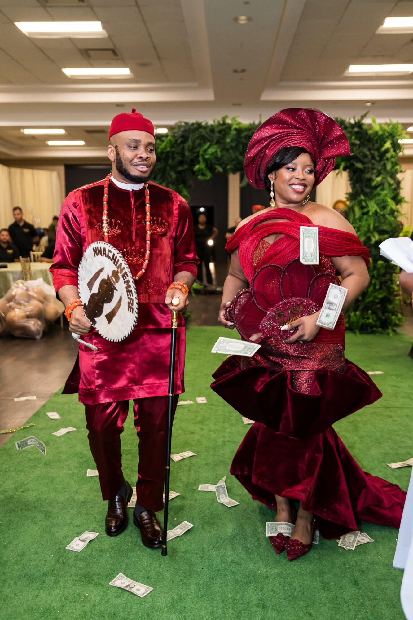 Two people dressed in traditional Nigerian attire, standing indoors with green plants behind them, holding fans and money on the floor.