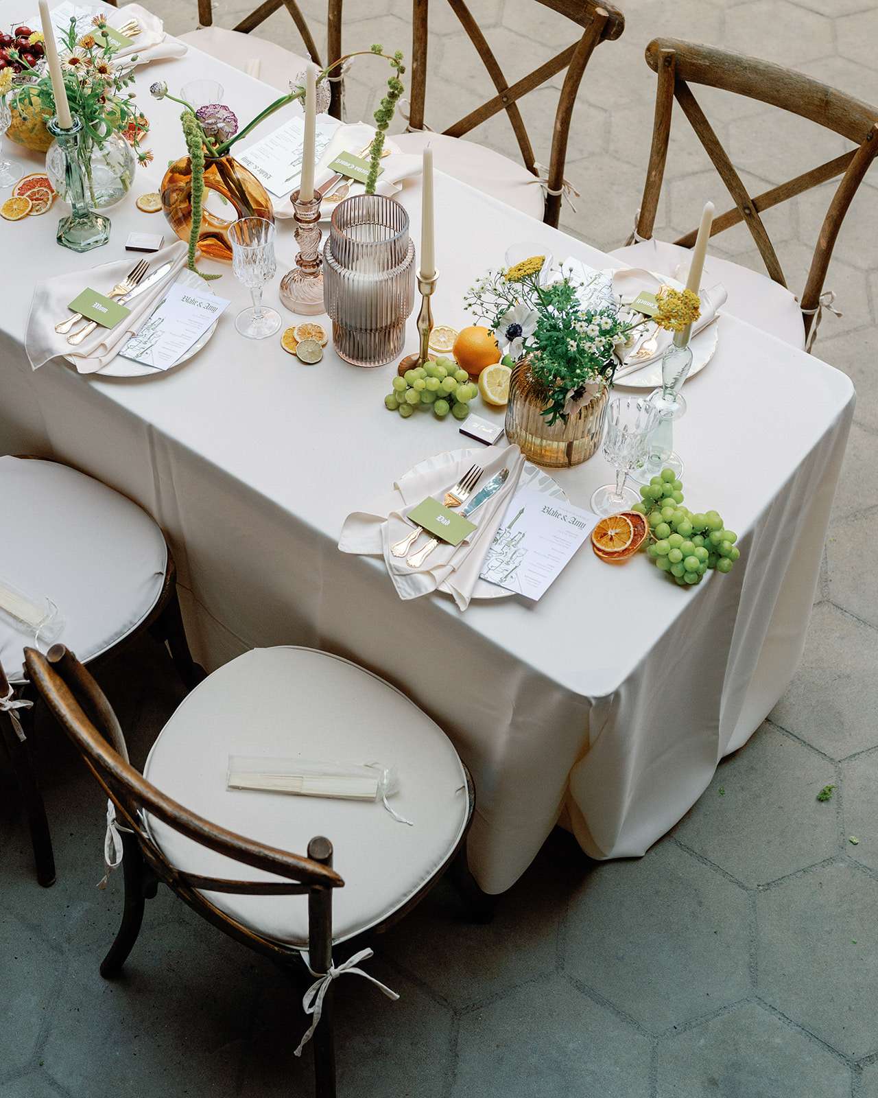 Table set with flowers, candles, fruits, and place settings, surrounded by wooden chairs with white cushions.