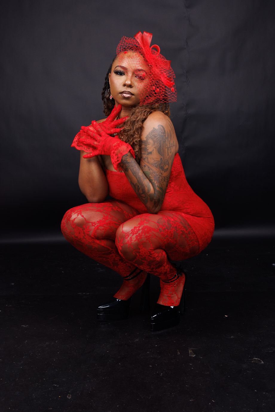 Person with red hair and tattoos crouching in a red outfit against a black background.