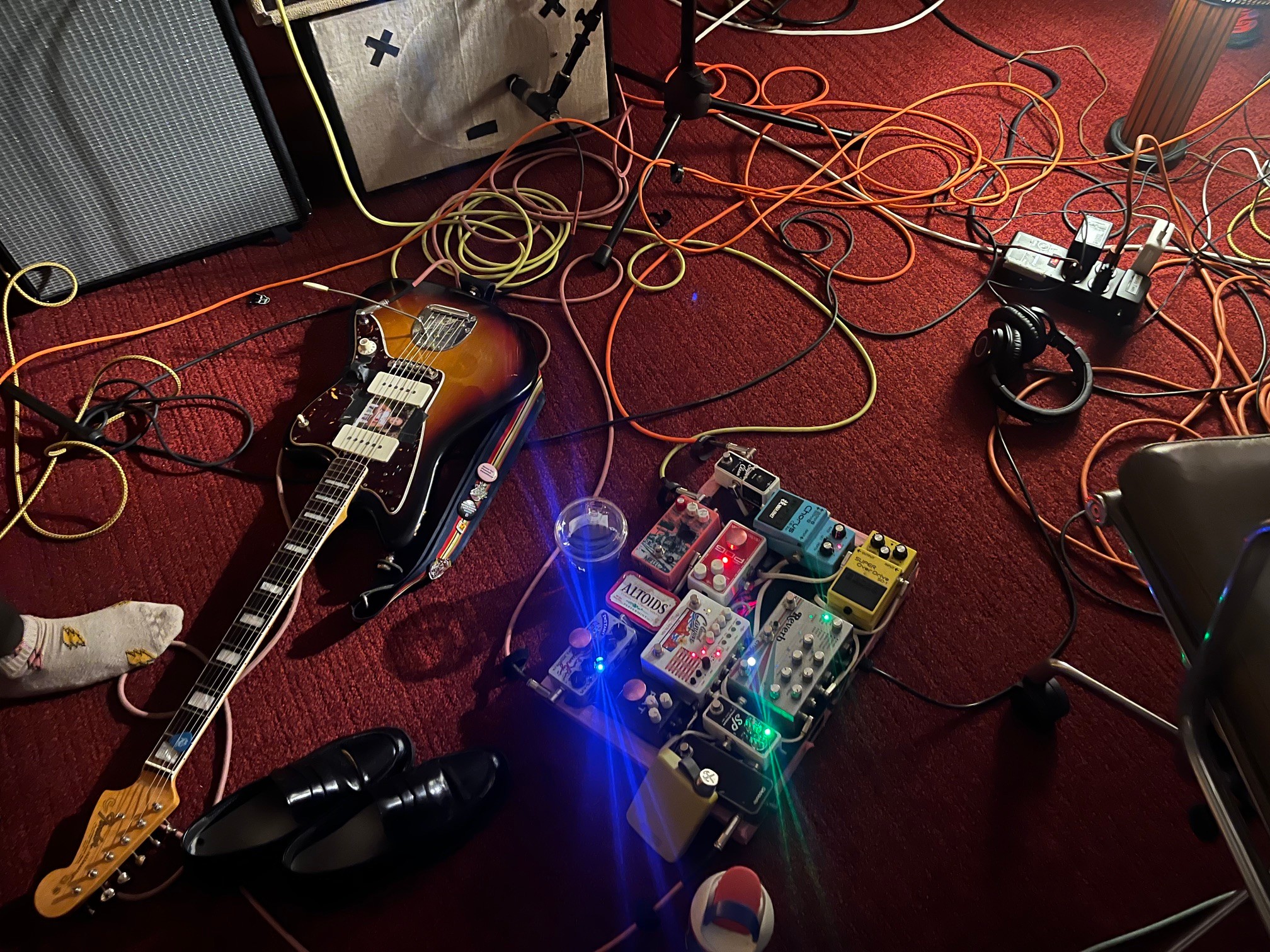 Guitar, pedals, and cables on a red carpeted floor with audio equipment and headphones, viewed from above.