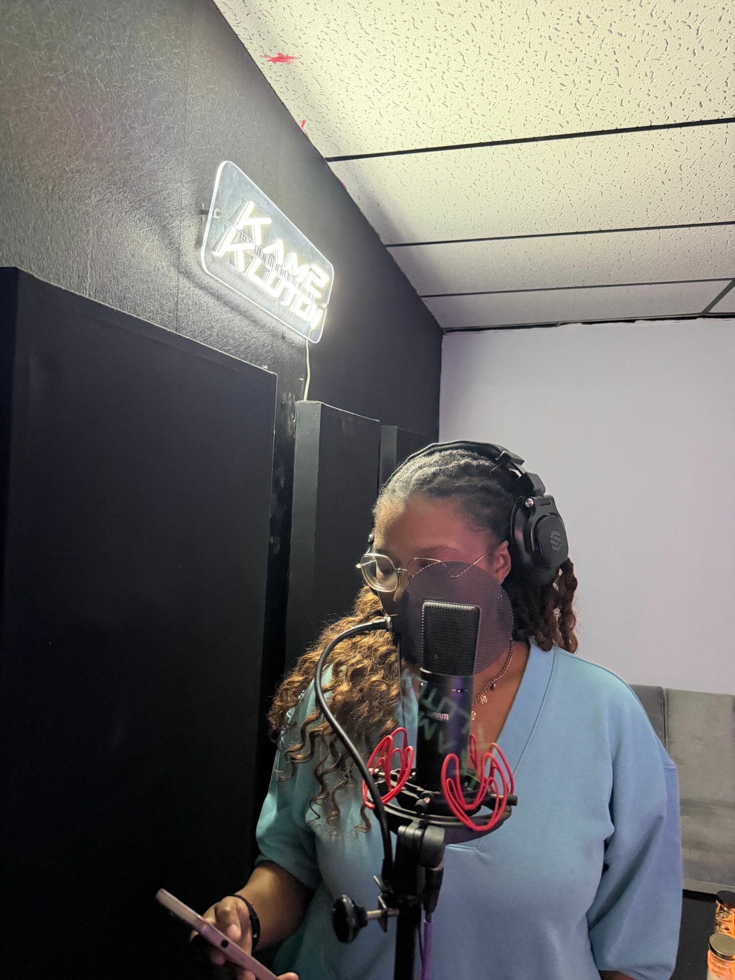 Person with headphones and face mask in a recording studio, holding a phone, with a sign on the wall, ceiling tiles, and black panels.