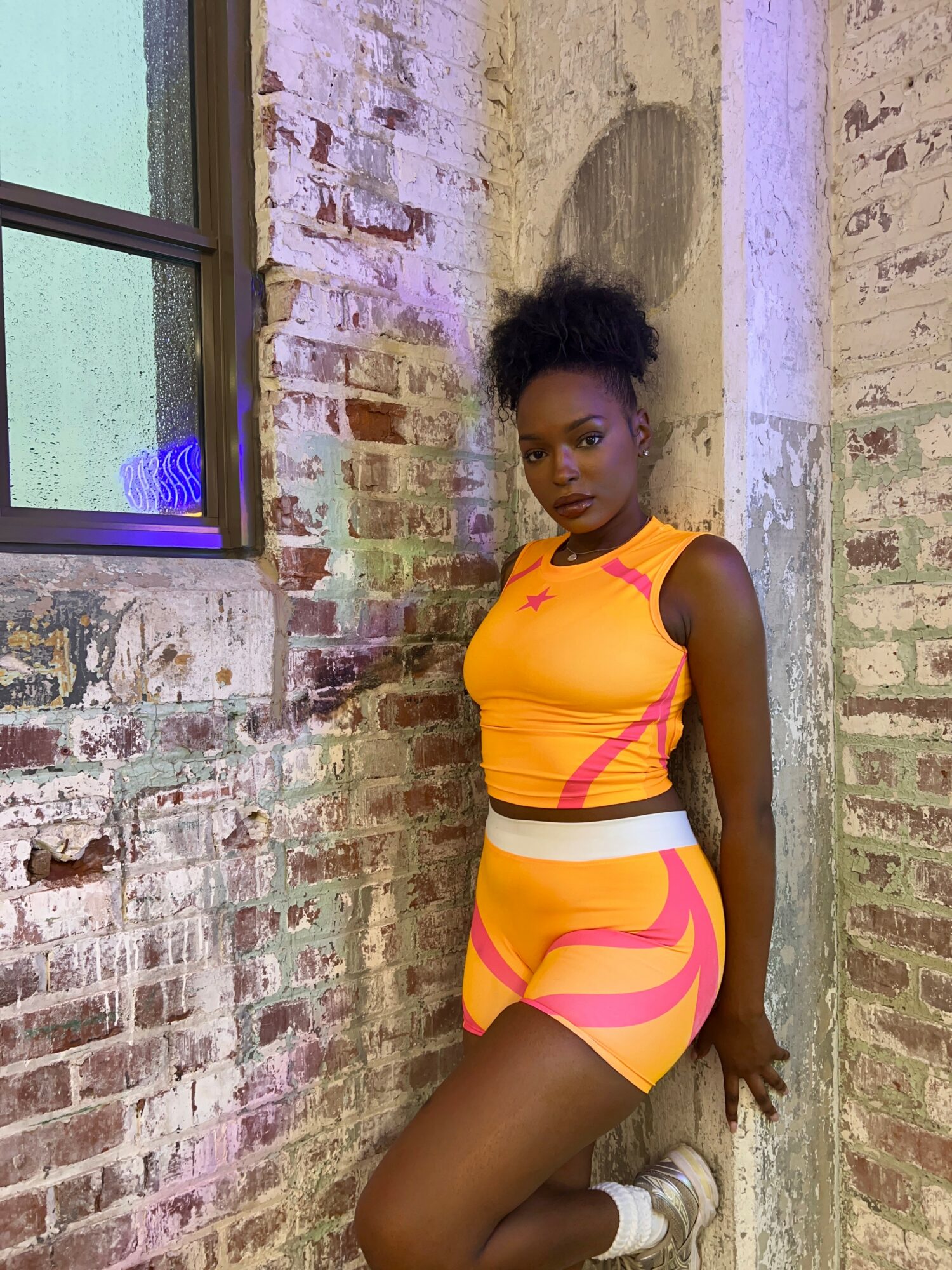 Young woman with natural hair in a yellow and pink athletic outfit standing against a brick wall.