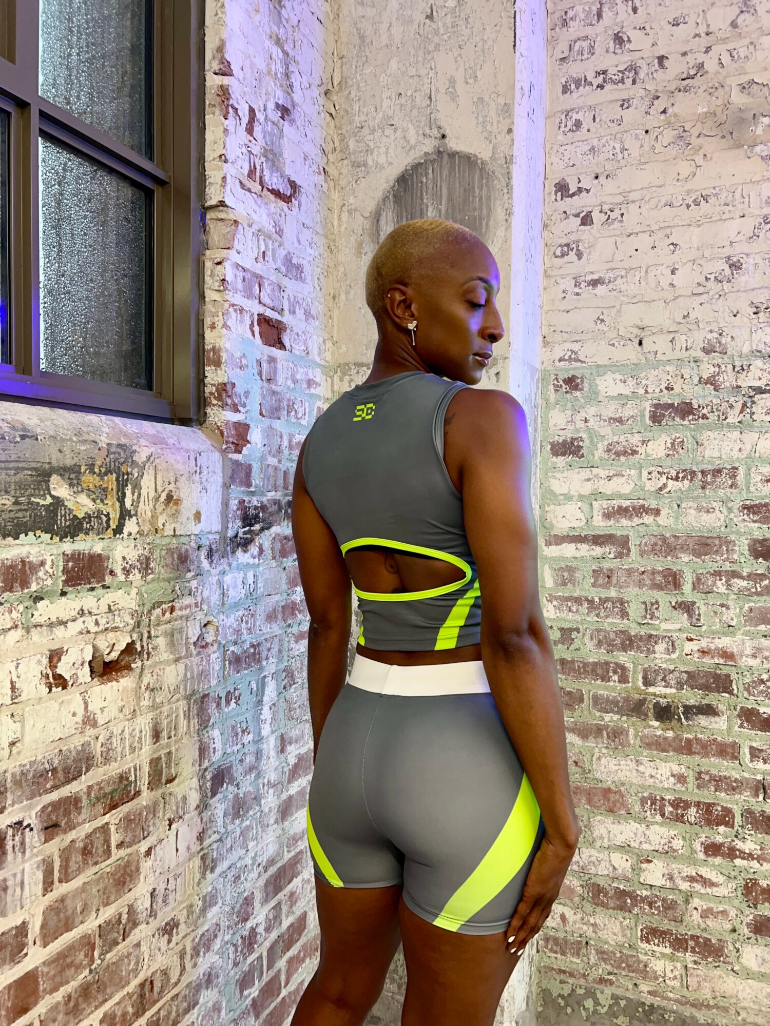 Person with short blonde hair in athletic wear standing against a brick wall, facing sideways with eyes closed.