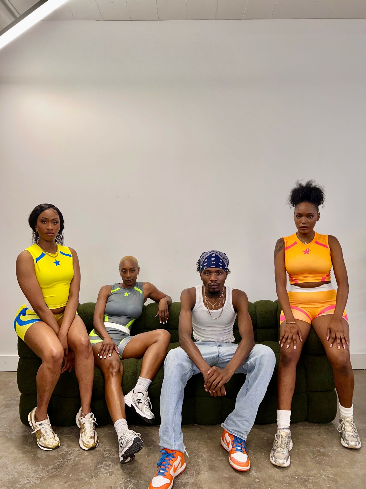 Four people sitting on a green couch against a plain white wall, wearing athletic clothing and sneakers.
