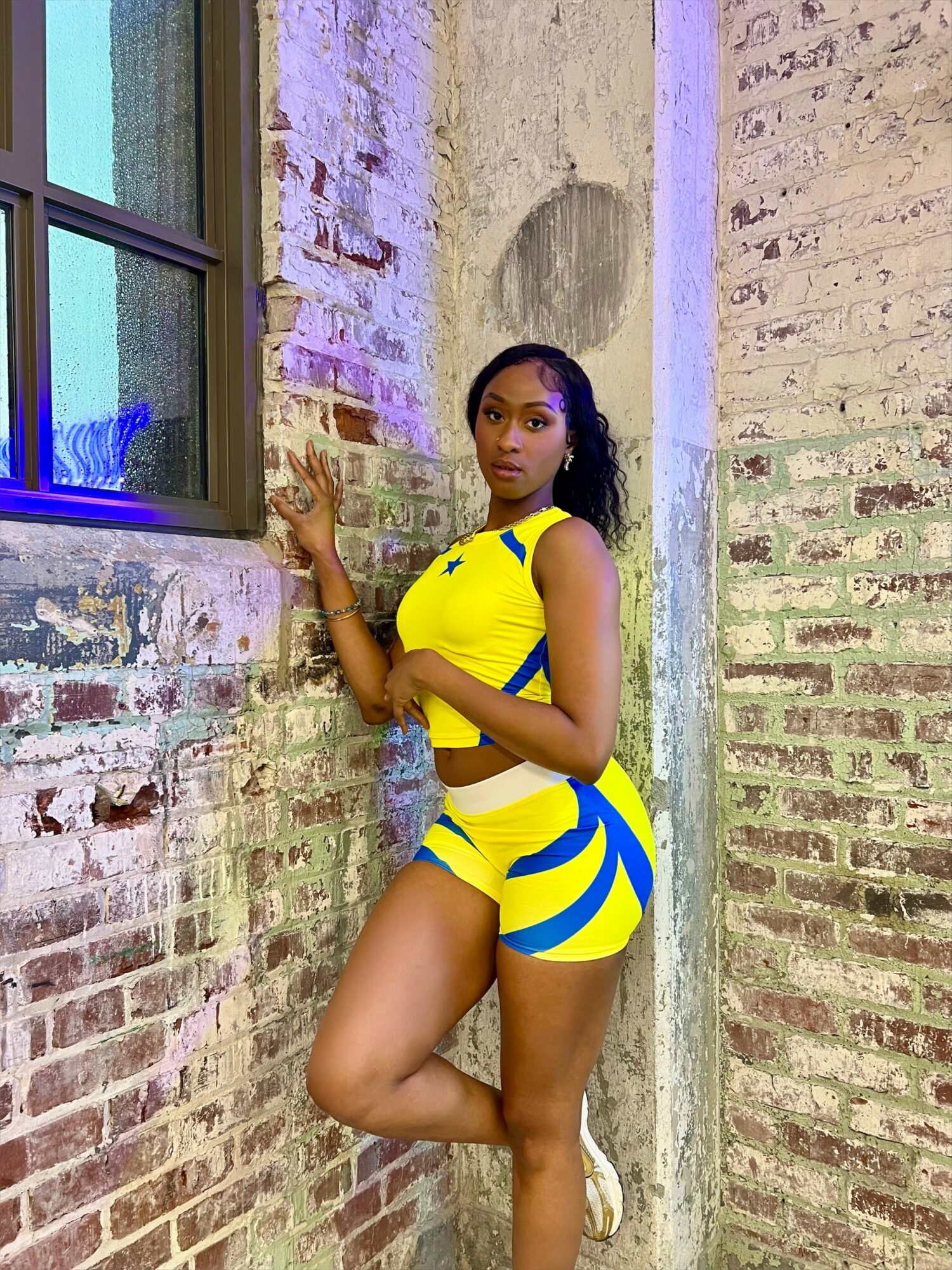 Woman in yellow and blue athletic outfit standing against a brick wall with a window, posing with one hand on the wall.