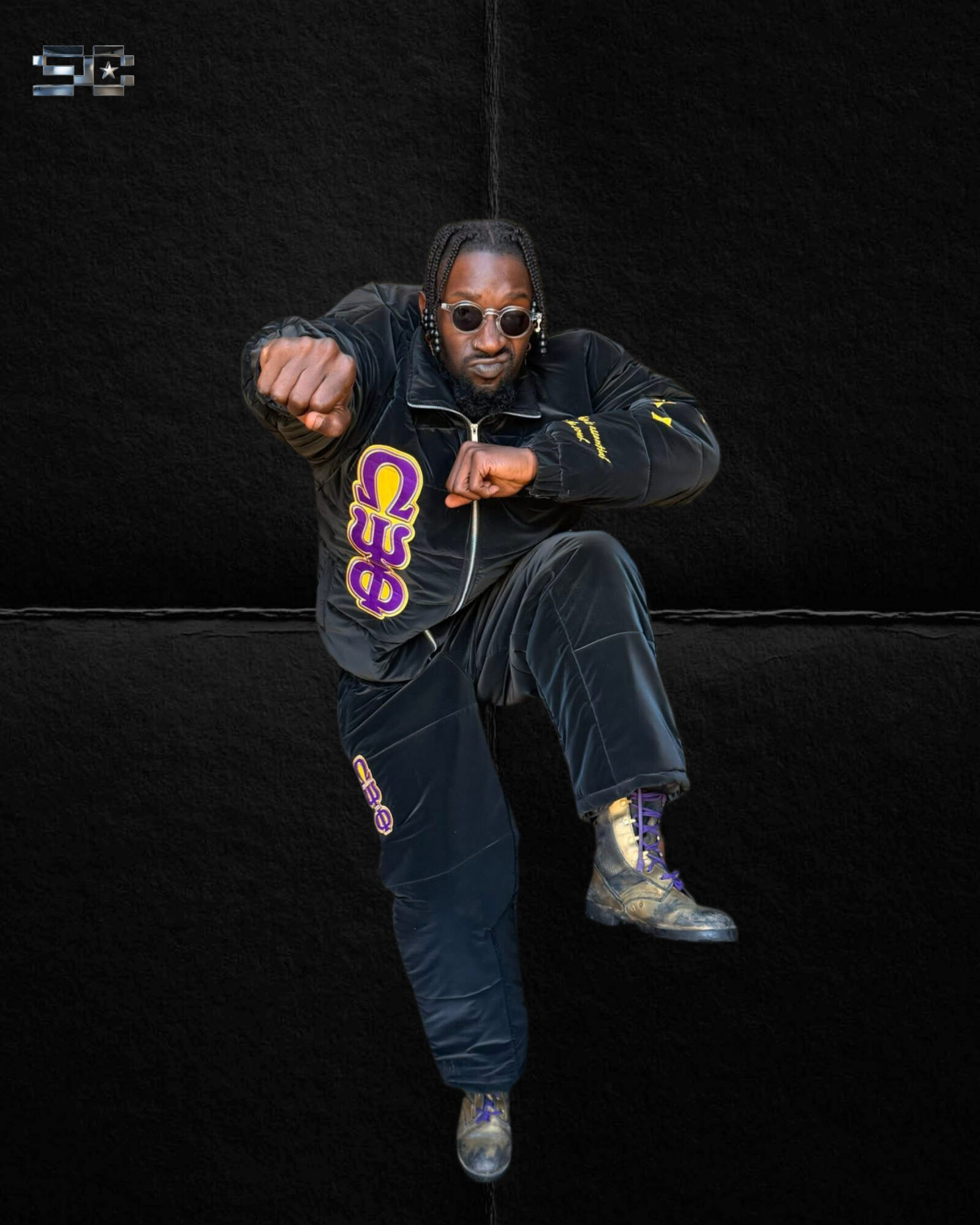 Person with sunglasses and dreadlocks poses with one leg raised, wearing a black jacket and pants, against a black background.