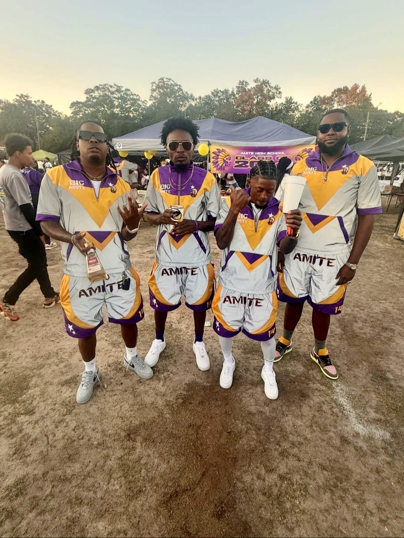 Four cricket players in purple, white, and yellow uniforms standing outdoors on a dirt field, with tents and trees in background.
