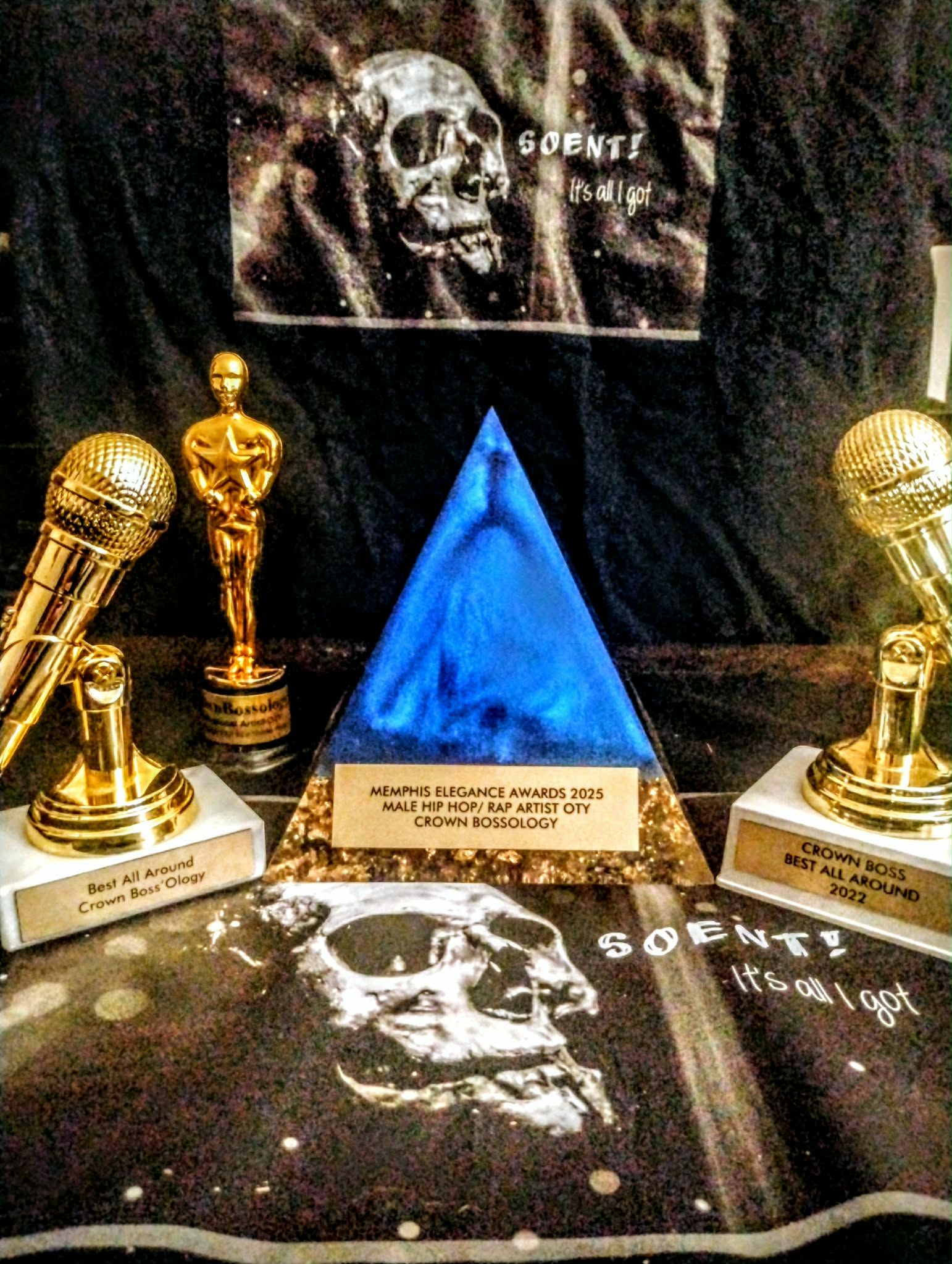 Awards and trophies displayed with a blue pyramid-shaped trophy in the center, surrounded by two gold microphone trophies, and posters with skull images.