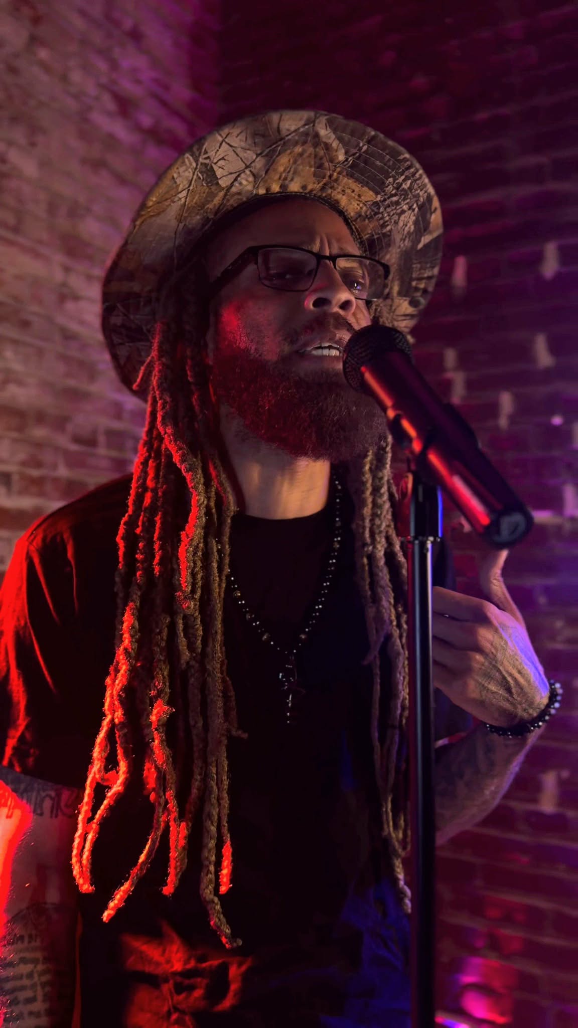 Person with long dreadlocks, wearing a hat and glasses, singing into a microphone on stage with purple lighting.