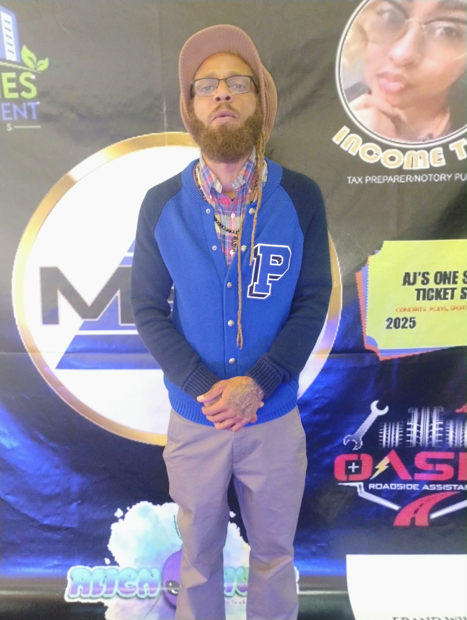 Man with beard, glasses, and a hat standing in front of a promotional backdrop, wearing a blue jacket and light-colored pants.
