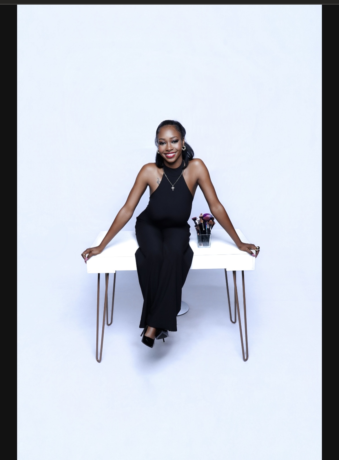 Woman in black dress sitting on a white table with a small flower arrangement, smiling, against a plain light background.