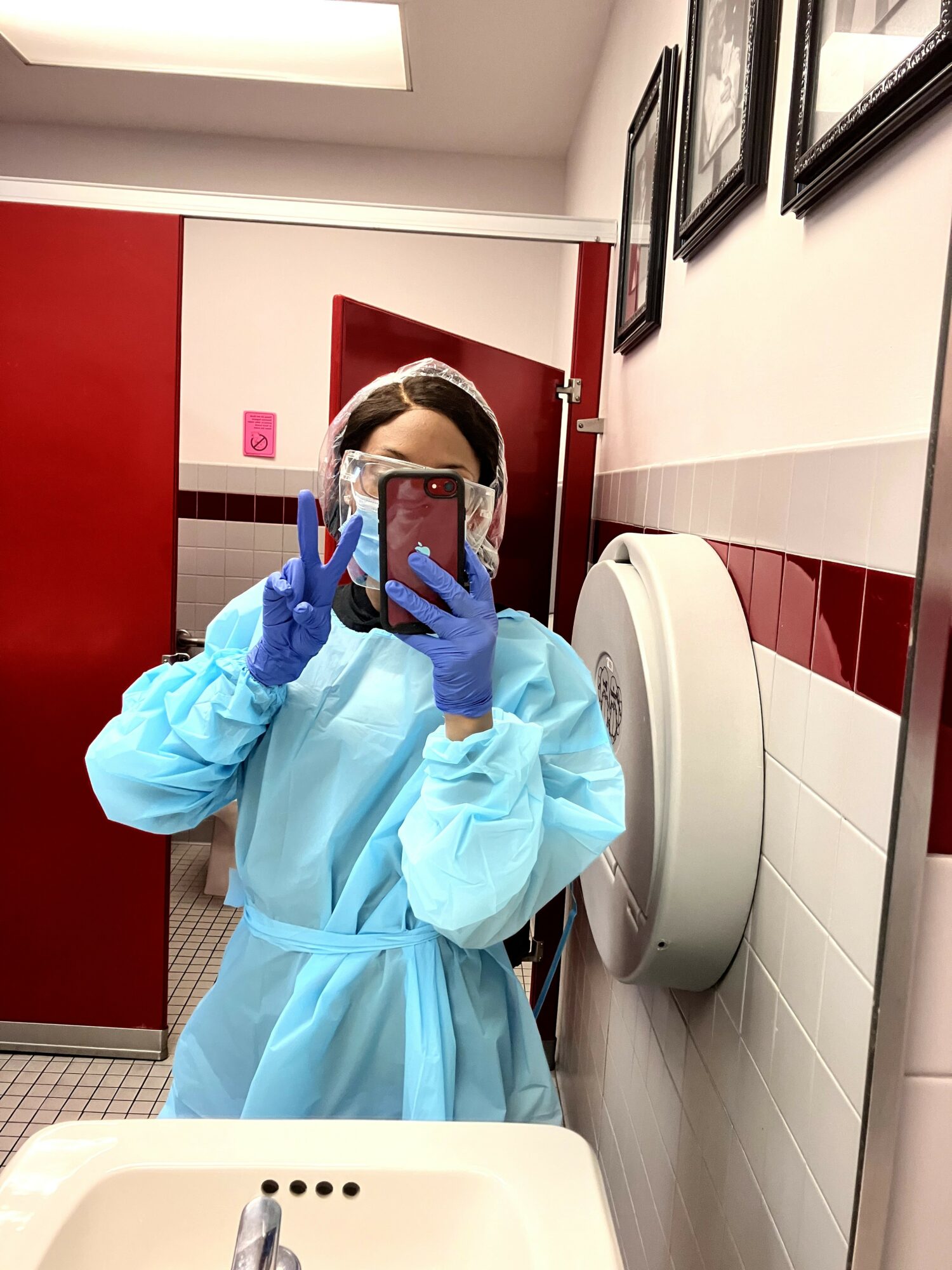 Person in protective gown and gloves taking a mirror selfie in a restroom.